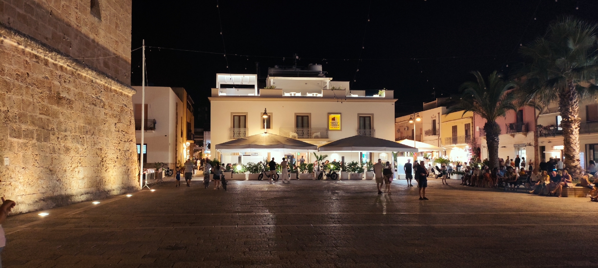 This is a wide-angle nighttime photograph of a paved plaza in San Vito Lo Capo, Italy, with buildings and people. On the left, a large, ancient stone building with illuminated rough-hewn blocks dominates the foreground. A white pole stands near its corner, and string lights are visible overhead. Across the plaza, a row of modern buildings with balconies and windows is illuminated. In the center of the plaza, there are several outdoor seating areas with umbrellas and plants, suggesting restaurants or cafes. Many people are seen walking, sitting, and standing throughout the plaza, some are in small groups, others individually. Two palm trees are visible on the right side of the image, with their fronds lit by external light sources. The ground is paved with cobblestones.