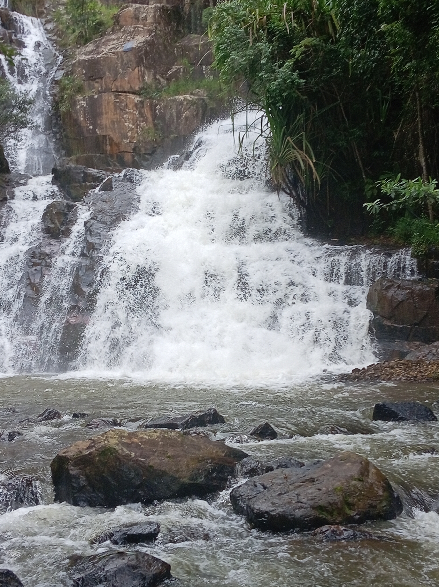 The image depicts a multi-tiered waterfall flowing over rocky terrain, with lush green vegetation, including bamboo, surrounding the upper section and sides. The water is turbulent and white, characteristic of a strong flow, cascading down into a rocky riverbed. Large, dark rocks are scattered throughout the river in the foreground. The overall scene suggests a natural, somewhat wild environment. This waterfall is located in the city of Da Lat, Vietnam, known for its numerous natural attractions. There are no people visible in the image.