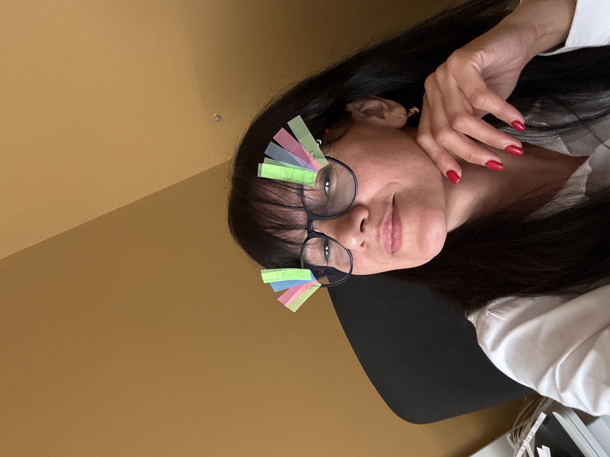 The image shows a woman with long, dark hair, wearing glasses with colorful paper strips attached to the frames. Her left hand is resting near her face, and she is wearing red nail polish. She is wearing a white shirt and appears to be sitting in a chair with a black back. The background is a plain, tan wall. The location is Unknown, Unknown.