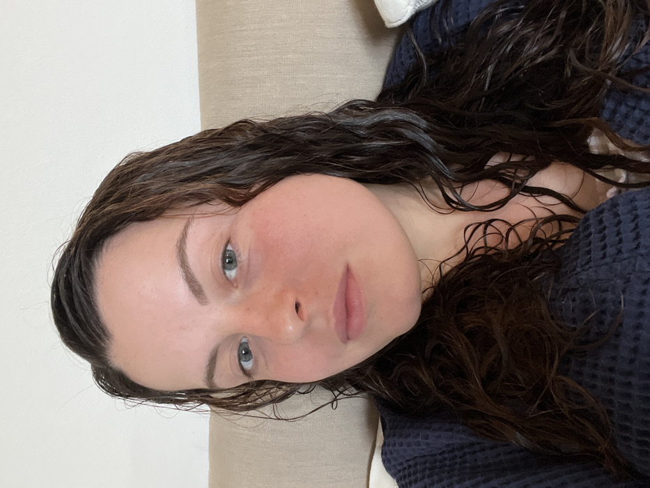 A close-up, head-and-shoulders portrait depicts a female subject in an indoor environment. The subject has fair skin, light-colored eyes (appears blue-grey), and dark brown, wavy hair that appears wet or damp, extending over her shoulders and chest. Her cheeks and nose exhibit a flushed appearance. She is looking directly at the camera with a neutral expression. She is wearing a dark blue, possibly navy, waffle-knit robe or similar garment, with a lighter-colored undergarment visible at the neckline. The background consists of a light-colored, textured fabric, likely a headboard or couch back, with a partial view of a white pillow to the right. The scene is located in Bad Sobernheim, Germany.