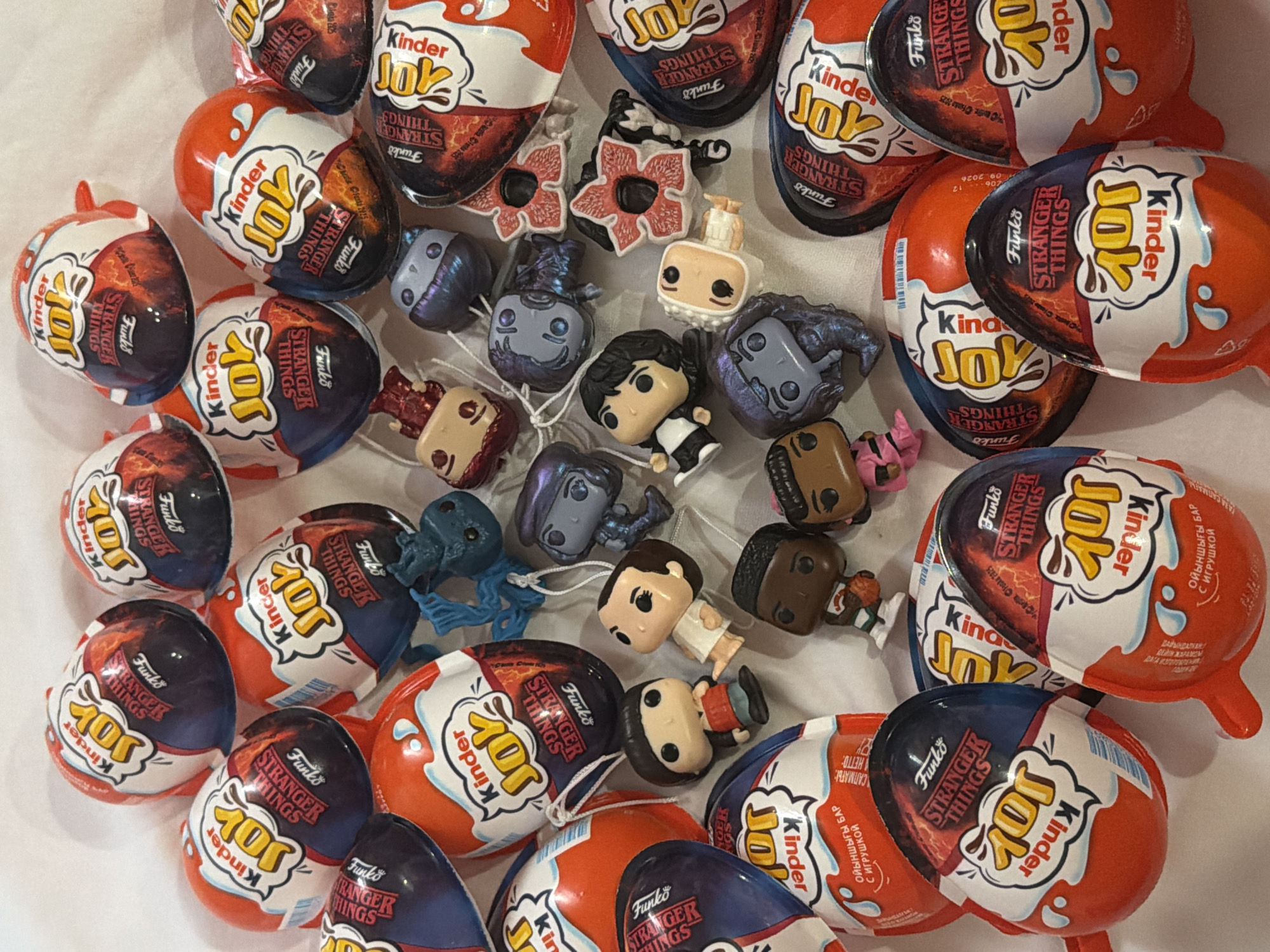 The image shows a collection of "Kinder Joy" eggs with "Stranger Things" branding, intermingled with small "Funko Pop!" figures of characters from the "Stranger Things" series. The eggs are primarily red and blue, with a white section displaying the "Kinder Joy" logo and "Stranger Things" imagery. The "Funko Pop!" figures are small, stylized versions of various characters. They are scattered among the "Kinder Joy" eggs. The background is a white surface. There is nothing in the image to suggest that this photo was taken in Perm, Russia.