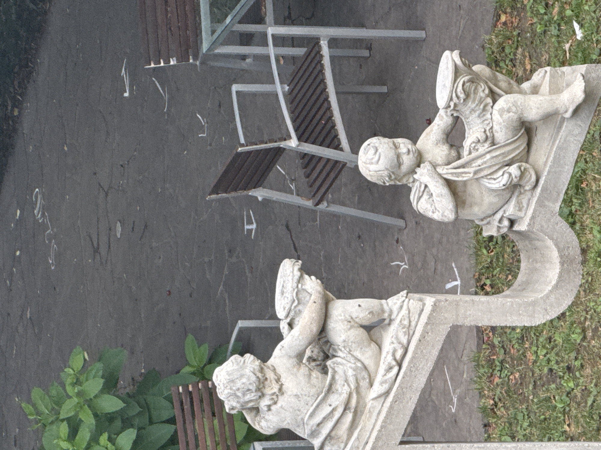 The image shows a stone statue of a cherub holding a decorative bowl, positioned on a stone structure resembling a musical note. In the background, there is a second, larger stone statue of a cherub holding a bowl, seated. Several outdoor metal chairs with wooden slats are visible, along with a glass-topped table. The ground appears to be asphalt with some white chalk markings. Green foliage and dry leaves are present on the ground around the statues and chairs. This scene is located in the courtyard of the Bratislava Castle in Bratislava, Slovakia.