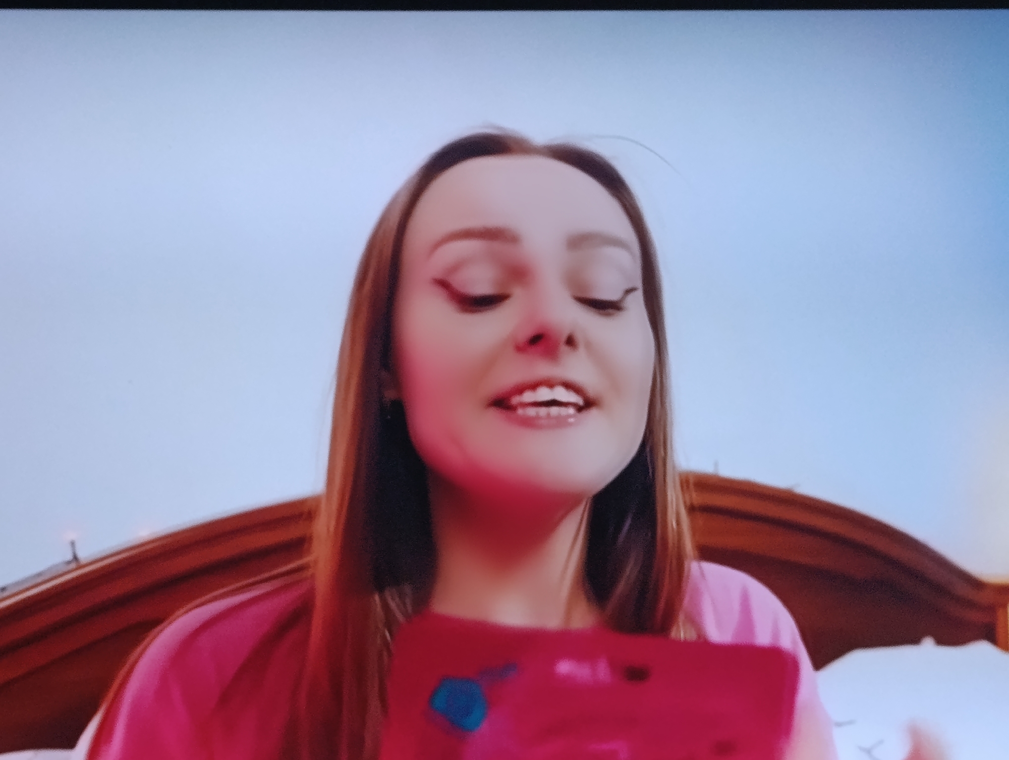 A young woman with long brown hair and prominent eyeliner is looking down at a pink object she is holding. She is wearing a pink shirt. The background is a light blue sky. The image is an indoor shot. No information is available about the location in Dnipro, Ukraine.