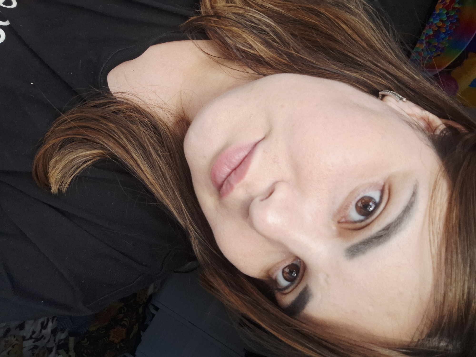 This is a close-up, head-and-shoulders, indoor portrait of a young woman. She has light skin, dark brown hair with lighter highlights, and brown eyes. She is wearing a black t-shirt and a small, silver hoop earring in her left ear. Her lips are painted a light pink. The background is dark and out of focus, with hints of colorful objects and patterned fabric. There is no indication of the specific location within Chaikovsky, Russia.