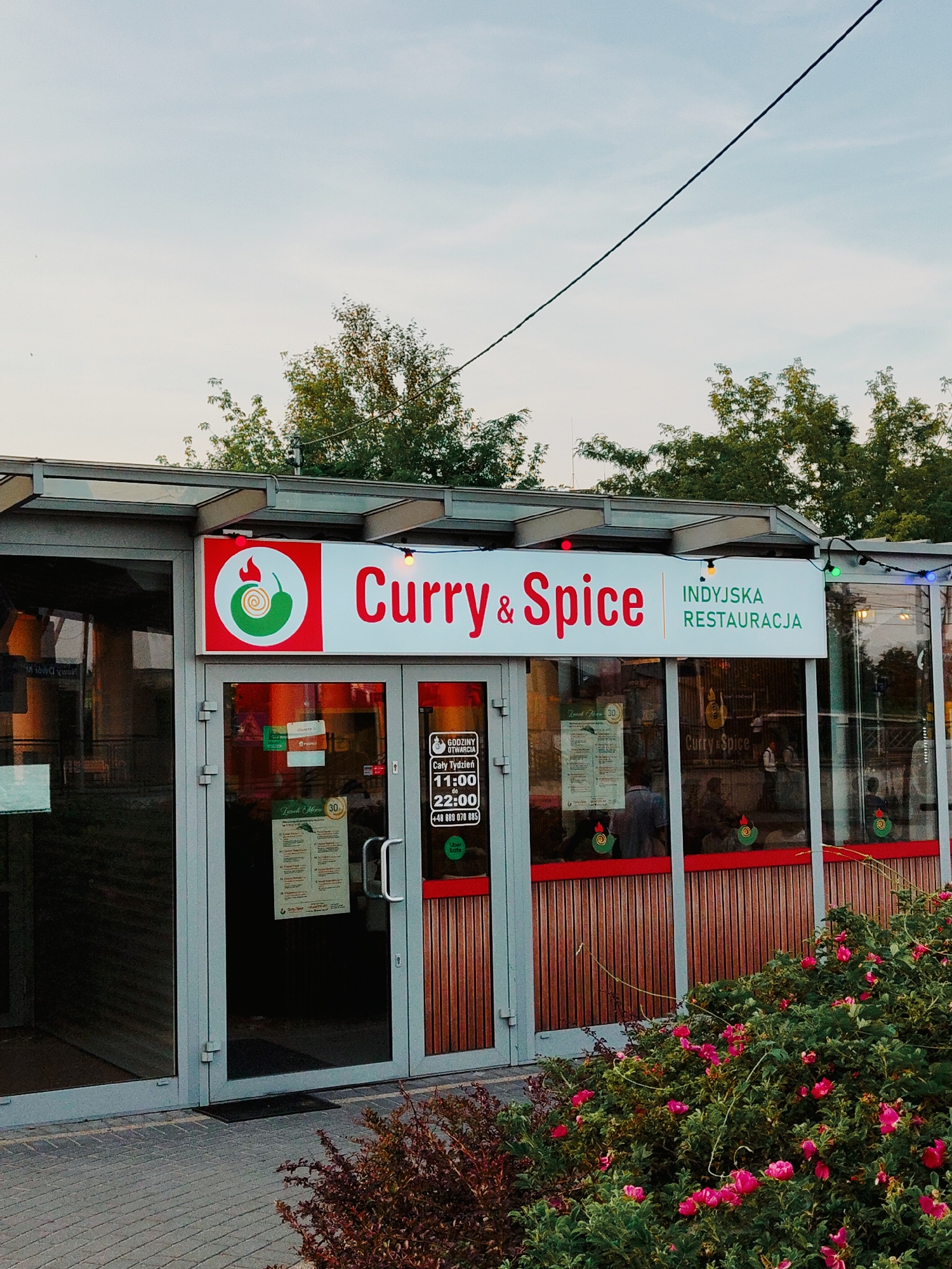 This media file features the exterior of an Indian restaurant named "Curry & Spice" in Nowy Dwór Mazowiecki, Poland. The restaurant has large glass doors and windows, with the name "Curry & Spice" prominently displayed in red lettering above the entrance, accompanied by a logo featuring a chili pepper and a spiral. To the right of the restaurant name, it is identified as an "INDYJSKA RESTAURACJA" (Indian Restaurant).
Visible on the glass doors are various signs, including operating hours ("GODZINY OTWARCIA"), stating the restaurant is open "Cały Tydzień" (All Week) from "11:00 do 22:00" (11:00 AM to 10:00 PM), and a phone number "+48 888 070 085". A "Lunch Menu" is also displayed. Inside the restaurant, through the windows, people can be seen seated at tables, suggesting the restaurant is open for business.
The setting appears to be outdoors, with greenery and flowering bushes in the foreground. The lighting suggests it is either early morning or late afternoon, with a soft, natural light. The sky is visible and appears clear. The overall scene conveys a casual and inviting atmosphere for dining.