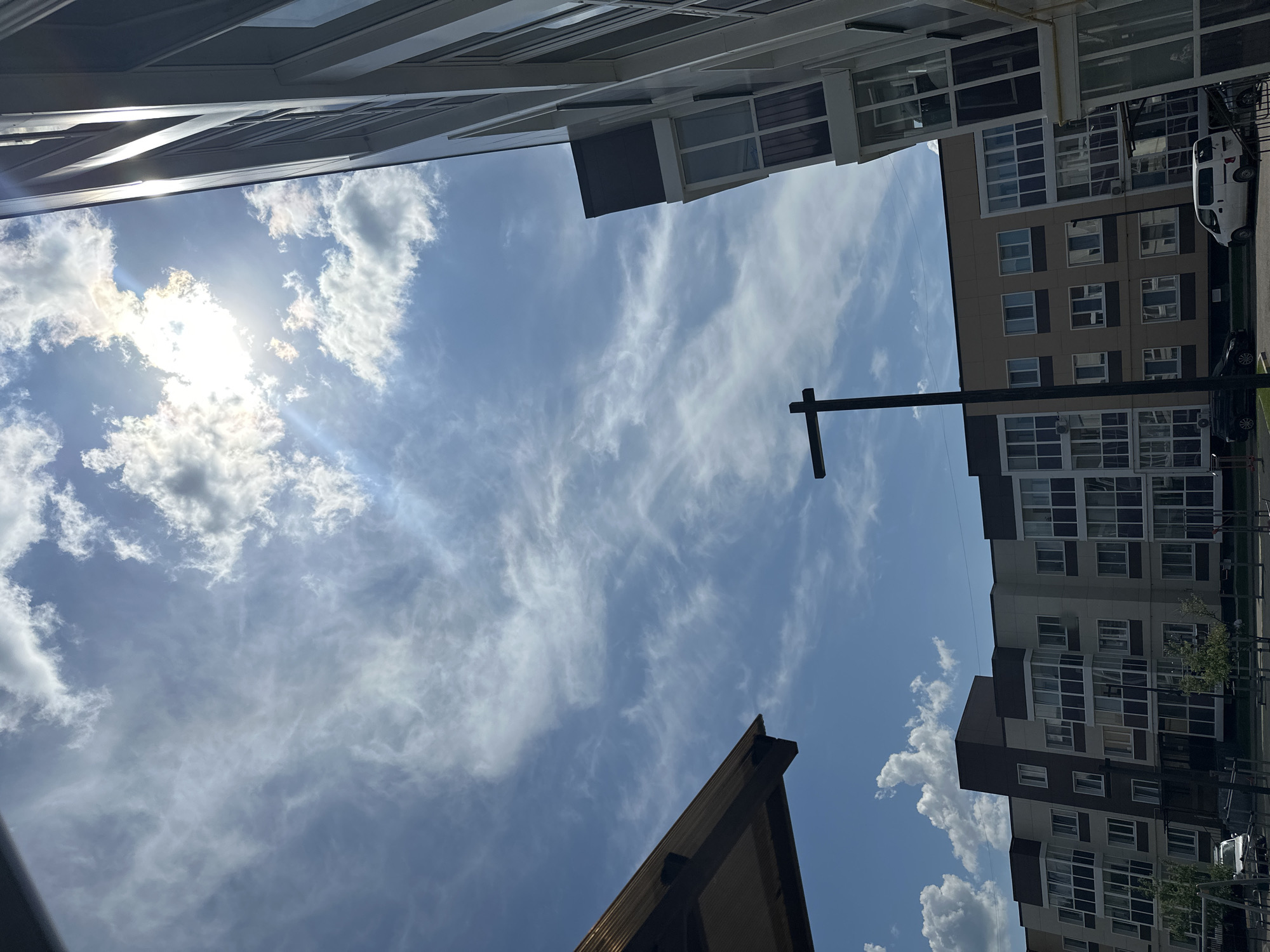 The image captures a street view in Ussuriysk, Russia, on a sunny day. The foreground is dominated by the right side of a modern, multi-story building with a facade of white vertical elements and glass balconies. To the left, the edge of a dark, angled roof structure is visible. In the middle ground, a street lamp with a black, T-shaped top stands against a backdrop of apartment buildings. These buildings are constructed with a combination of beige and dark brown sections and feature numerous windows and balconies. The sky above is a bright blue, streaked with thin, wispy clouds and illuminated by the sun, which is partially obscured by clouds in the upper right quadrant, creating a lens flare effect. In the lower portion of the image, a white van is parked, and hints of other vehicles and playground equipment are visible on the ground. The scene depicts an urban residential area.