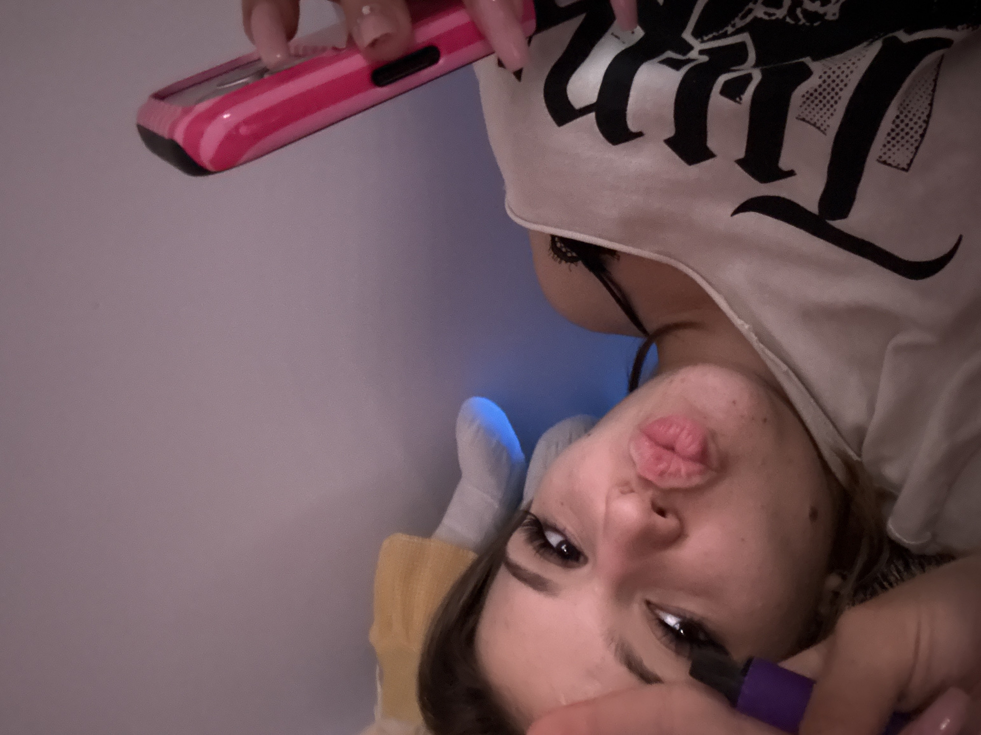 This image is an upside-down close-up shot of a young woman lying down and taking a selfie. She has light skin, dark hair, and full lips puckered into a "kissing" pose. Her right eye, with dark eyeliner and long eyelashes, is visible and looking towards the camera. She appears to be wearing a light-colored top with black lettering, possibly a graphic tee, and what looks like a black lace bra strap is visible beneath her shirt collar.
In her right hand, positioned above her head, she holds a pink and white striped mobile phone, which she is using to take the picture. Her fingernails are painted a light pink. In her left hand, which is closer to the viewer, she holds a purple object, possibly a marker or another small electronic device.
The background is a solid, light grayish-purple color, suggesting an indoor setting with plain walls. There's a subtle blue glow coming from behind her head, possibly from a light source or an electronic device. Part of a yellow fabric, perhaps a pillow or blanket, is visible beneath her head, and white socks are partially visible on her feet in the upper left corner of the frame. The time of day cannot be determined from the image, but the lighting suggests an indoor setting, possibly at night or with artificial light. The image conveys a playful and relaxed mood. No specific text is clearly legible on her shirt.