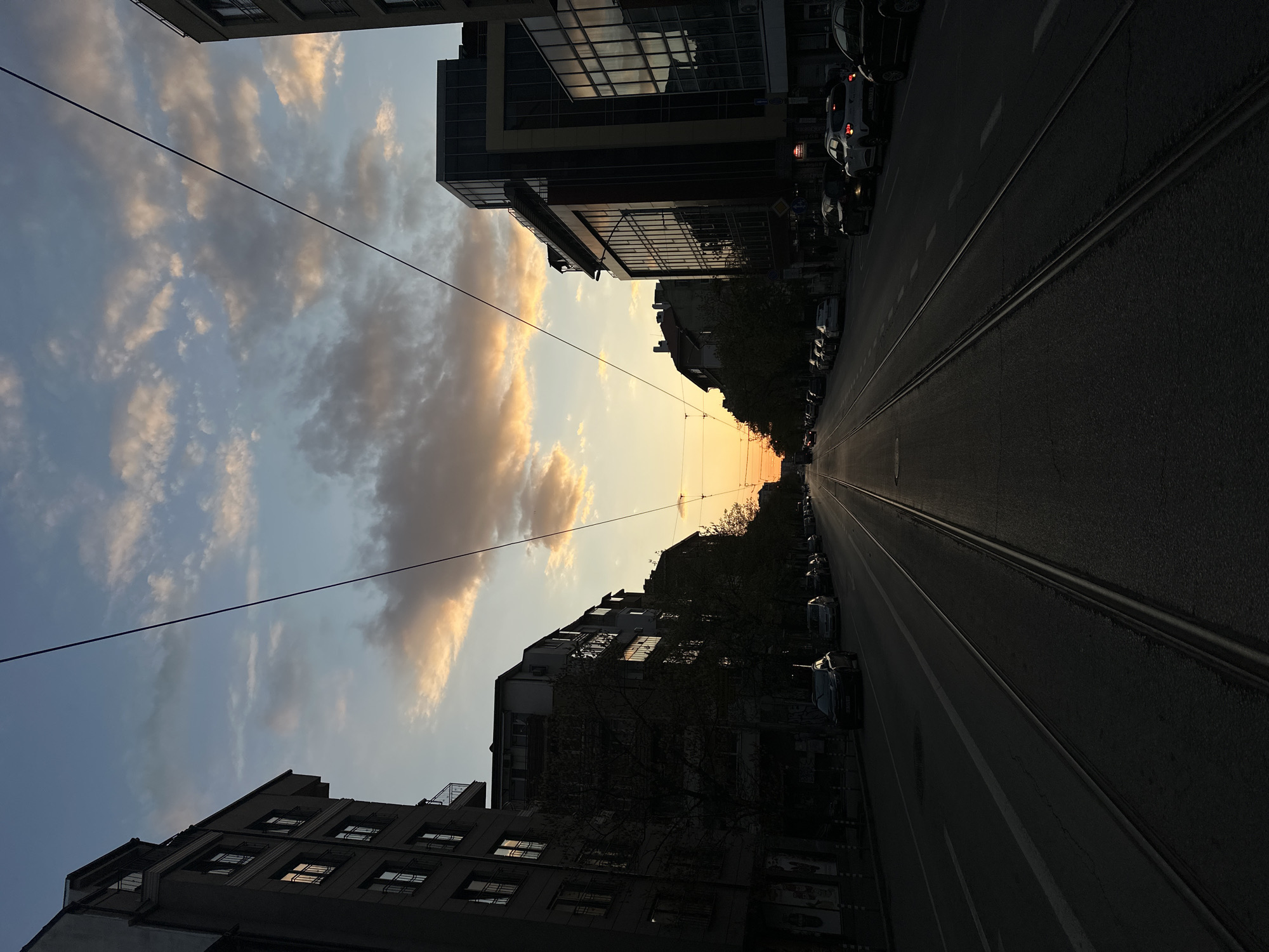 The image displays a city street in Sofia, Bulgaria, captured during either sunrise or sunset, looking down a straight road. The sky dominates the upper portion, featuring clear blue areas interspersed with scattered, wispy clouds that transition into vibrant golden and orange hues towards the low horizon, indicating the sun's position. Overhead utility lines are visible, stretching across the sky.
Tall buildings line both sides of the street, largely appearing as dark silhouettes against the bright sky. On the left side, a multi-story building with numerous windows reflects some of the sky's light. On the right, a more modern structure with a reflective glass facade is prominent, alongside other buildings and silhouetted trees.
The asphalt street features two sets of tram tracks running parallel down its center, receding into the distance and providing a strong leading line. Numerous cars are parked closely along both curbs of the street. The overall scene suggests a quiet moment in the city, illuminated by the warm, low light, with a peaceful atmosphere. While some indistinct signs are present on buildings, any visible text is unreadable due to distance and blur. No people or specific activities are discernible, showcasing a typical urban environment during a transitional period of the day.