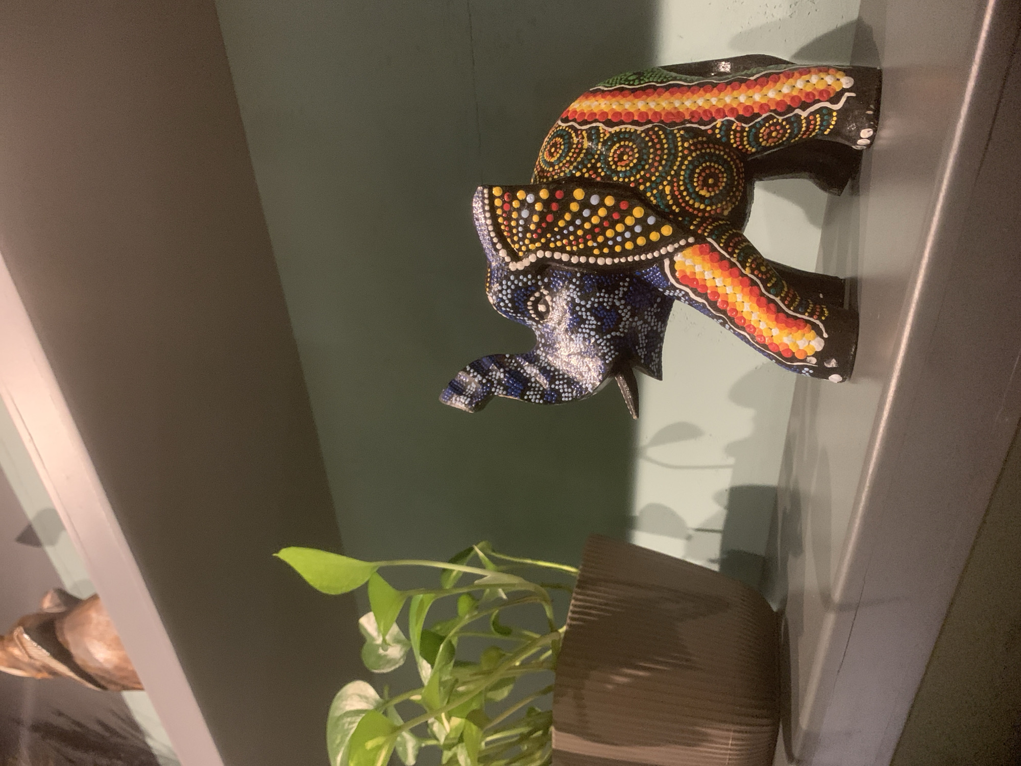 The image displays an indoor scene featuring decorative objects on what appear to be shelves, against a muted green wall. The photograph is oriented vertically, with the primary subjects appearing to be positioned on a vertical surface.
The most prominent subjects are two vibrantly decorated elephant figurines, situated in the upper right portion of the frame. The upper elephant features intricate dot art patterns in warm tones of red, orange, and yellow, contrasted with black. The lower elephant showcases similar dot art, but in cool tones of blue, white, and black. Both elephants have a stylized, textured appearance and small white tusks.
Below the elephants, towards the left and center of the frame, is a potted plant with numerous bright green leaves. The plant sits in a brown pot with visible vertical ribbing. A less distinct, wooden-colored object is partially visible on a shelf edge in the far left of the image.
The background consists of plain, light green walls. Shadows cast by the objects are visible on the wall, suggesting an internal light source. White or light-colored shelf edges are present on both the left and right sides of the image, reinforcing the setting of items displayed on shelves. No people or visible text are present in the image. The scene conveys a sense of home decor or an art display.