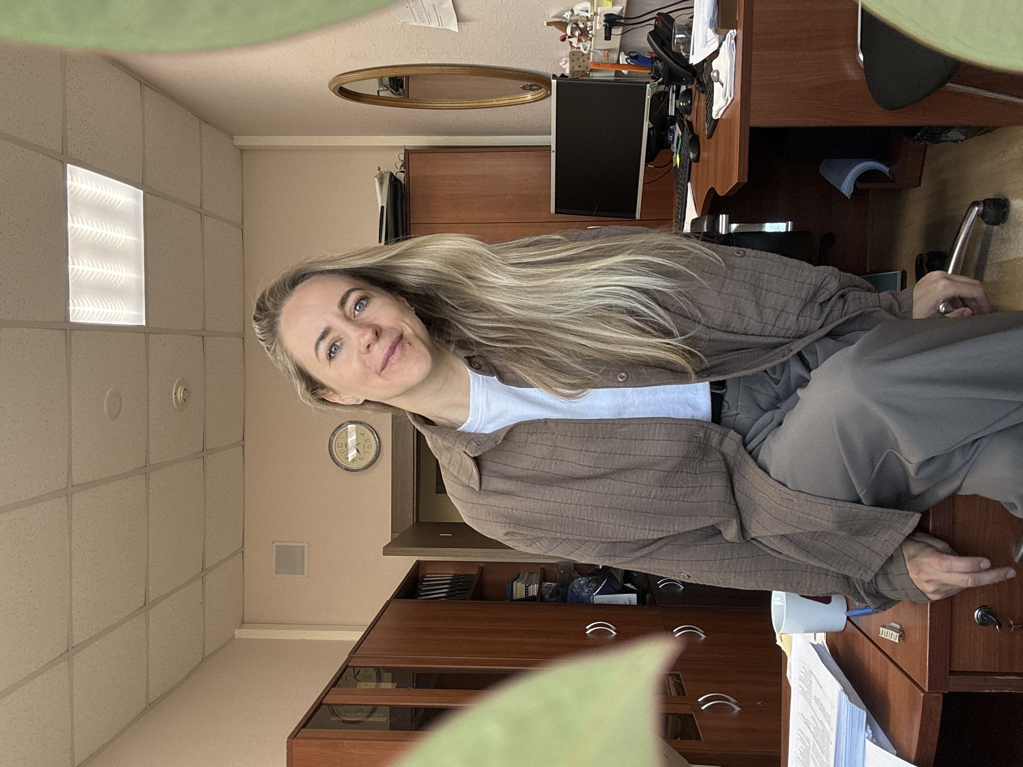 A woman is seated at a wooden office desk in what appears to be an office space. She has long blonde hair and wears a brown button-down shirt over a white t-shirt and grey pants. Her right hand rests on the edge of the desk, near a stack of papers and a white mug with a pen inside.
Behind her are wooden cabinets and a desk with a computer monitor and telephone. A gold-framed oval mirror is visible on the wall above the cabinets. A clock hangs on the wall, and the room is lit by fluorescent ceiling lights.
It is not possible to definitively identify the location as Chaikovsky, Russia, from the image alone.