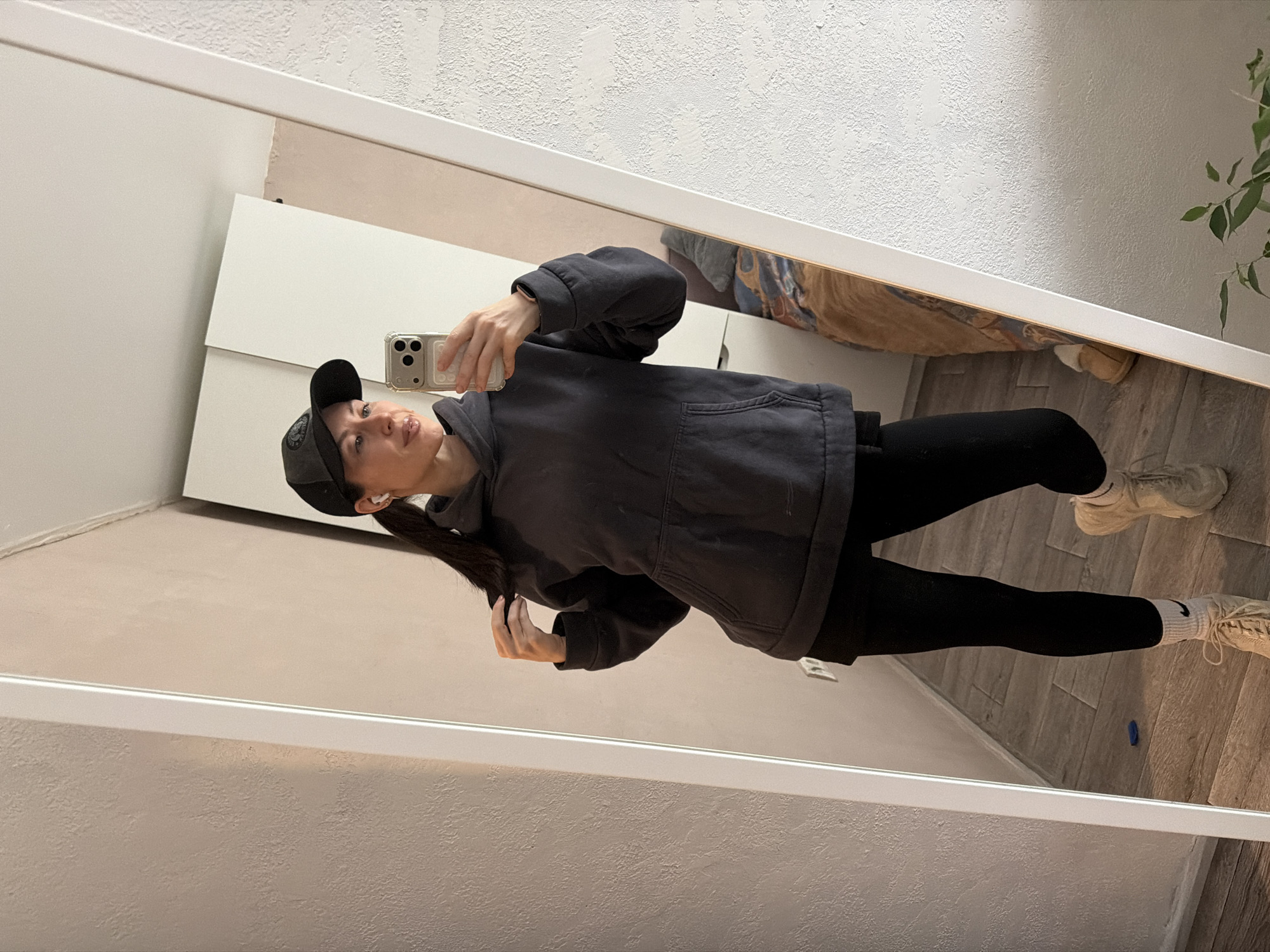 A woman is taking a mirror selfie indoors in what appears to be a room in Perm, Russia. She is depicted from the waist up, holding a light-colored smartphone with a triple-camera system in her right hand, pointed towards her reflection. She is wearing a dark baseball cap, a dark grey or black oversized hoodie, black leggings, light-colored sneakers, and white socks that feature a visible black Nike checkmark logo. Her long, dark hair is pulled back, and her left hand is positioned near her hair, possibly adjusting it. She has a neutral expression, looking towards the camera or her reflection.
The mirror reflects parts of the room, including a white door or cabinet on the left, a textured white ceiling, and light wooden or laminate flooring. A small blue object is visible on the floor near her reflected feet. A green potted plant is partially reflected in the upper right corner of the mirror. The lighting is even, suggesting an indoor setting. The logo on her baseball cap is circular and indistinct, with text that is too blurry to read clearly.