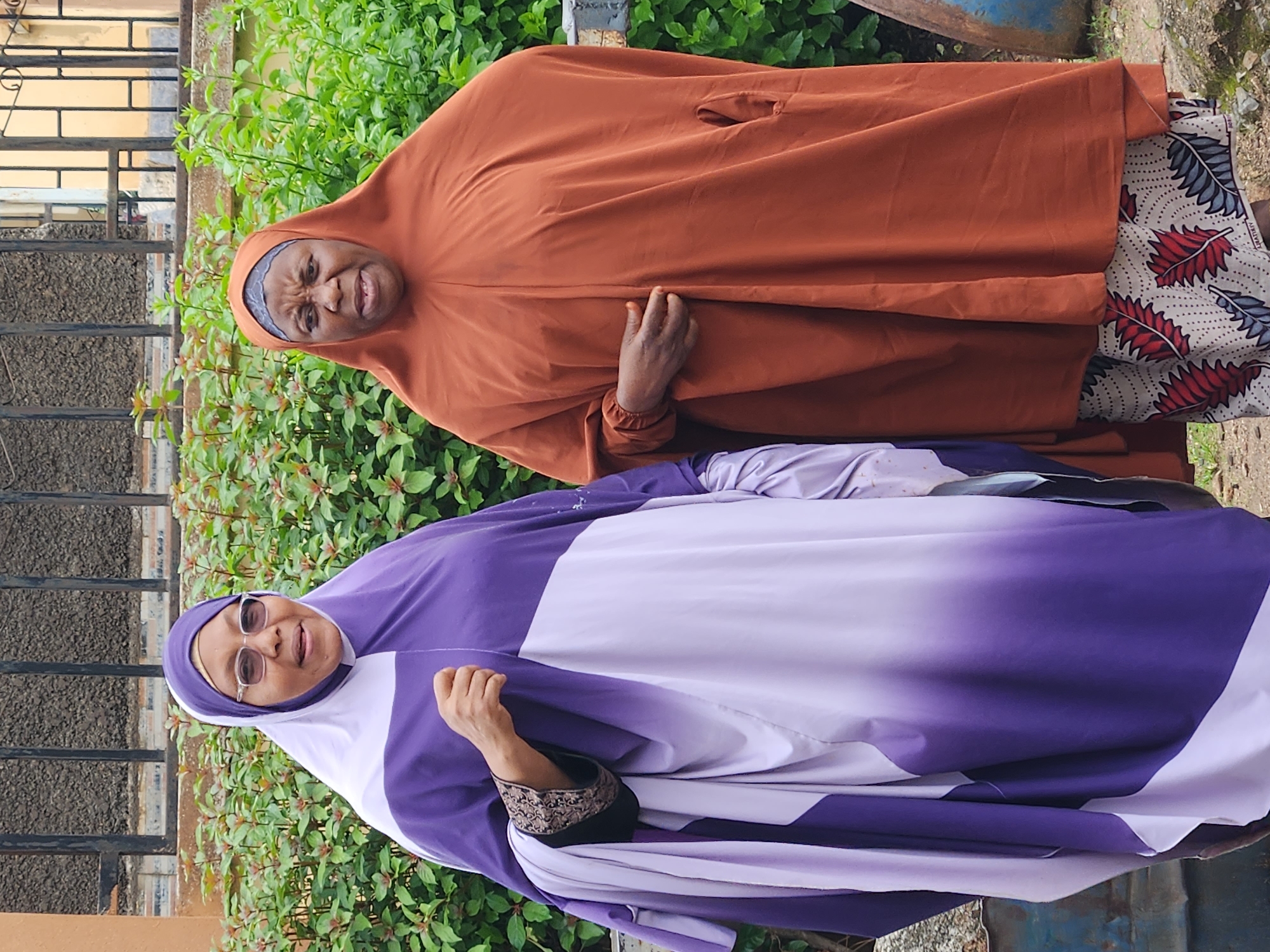 Two women are standing outdoors in front of a hedge and a fence. The woman on the left is wearing a purple and white hijab and a matching robe with decorative black trim on the sleeves. She is wearing glasses and smiling. The woman on the right is wearing an orange hijab and robe over a patterned skirt. She is looking at the camera with a concerned expression. The background consists of lush green foliage and a dark metal fence with yellow accents. There are also some industrial objects visible to the left and right of the women, possibly barrels or bins. The ground appears to be dirt and small stones.