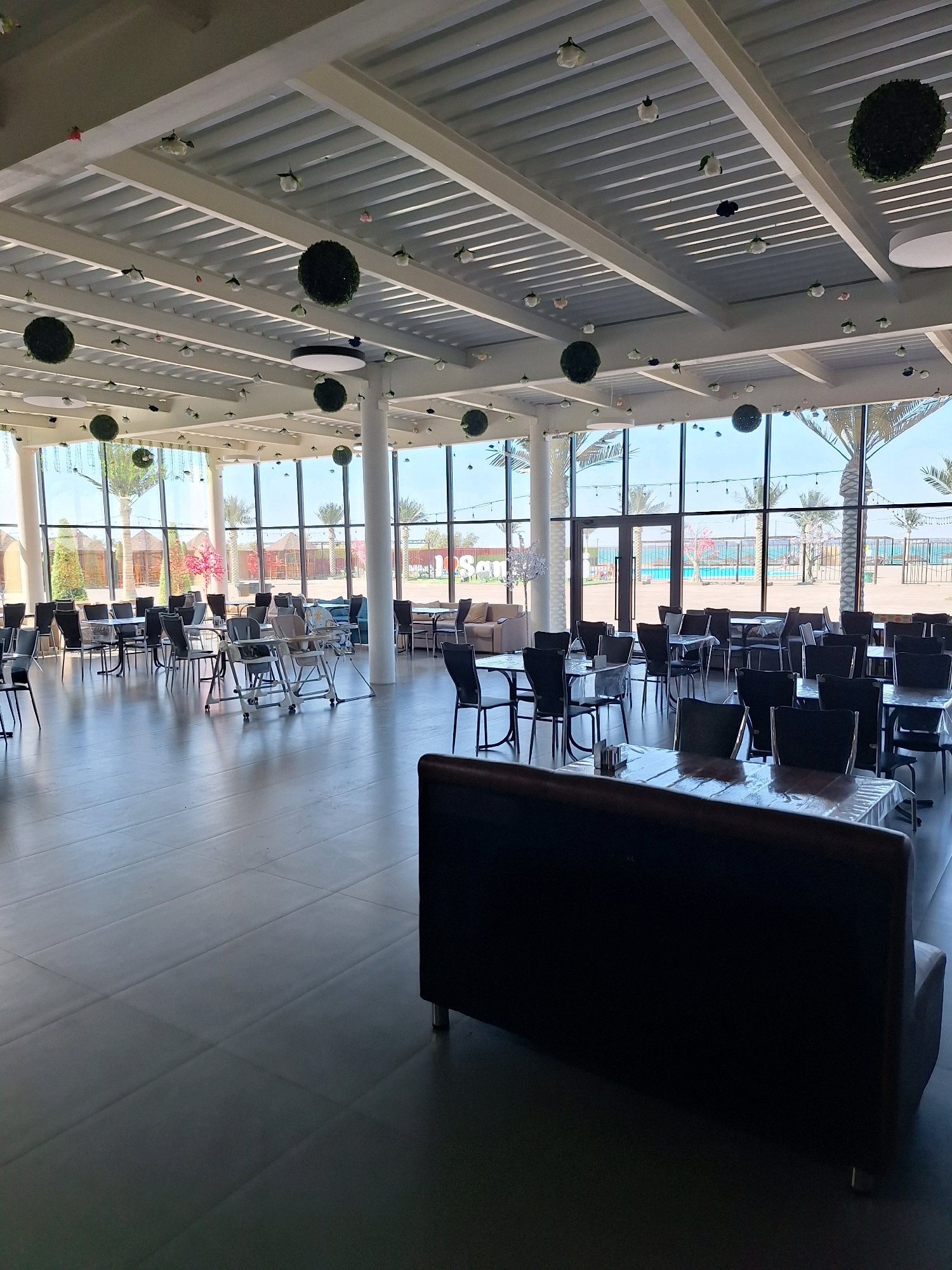 The image shows a spacious indoor seating area with large windows overlooking an outdoor pool and palm trees, indicative of a resort or entertainment complex. The room is furnished with numerous tables and chairs, some of which are covered with transparent plastic. There are also several high chairs, suggesting a family-friendly environment. Decorative spherical moss balls hang from the ceiling, along with small artificial flowers. The flooring is a dark, tiled material. The overall atmosphere is bright and open, with natural light streaming in through the windows. The scene appears to be unoccupied at the time the photograph was taken. Based on the context provided, this location is identified as the "Los Santos" cafe within the Balkhash Bay resort in Balkhash, Kazakhstan.