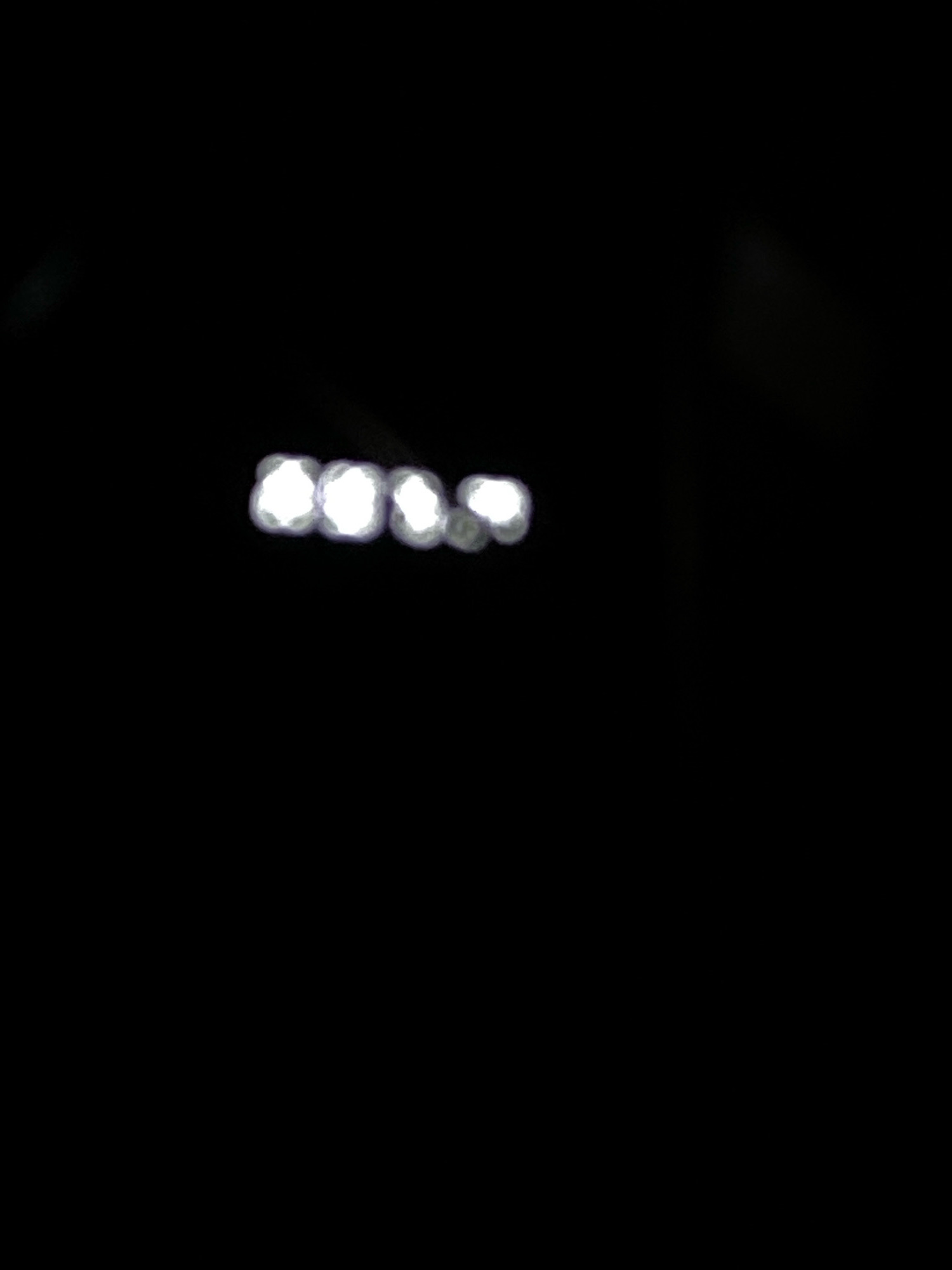 The image shows a blurry representation of lights, possibly from a vehicle or streetlights, against a dark background. The lights appear as circular or hexagonal shapes with a bright white center and softer edges, suggesting they are out of focus. The arrangement of the lights is horizontal, with several distinct illuminated points. There are no discernible people, objects, actions, or interactions visible in the image due to the blurriness and darkness. The location within Klurak, Indonesia, cannot be determined from the visual information provided.