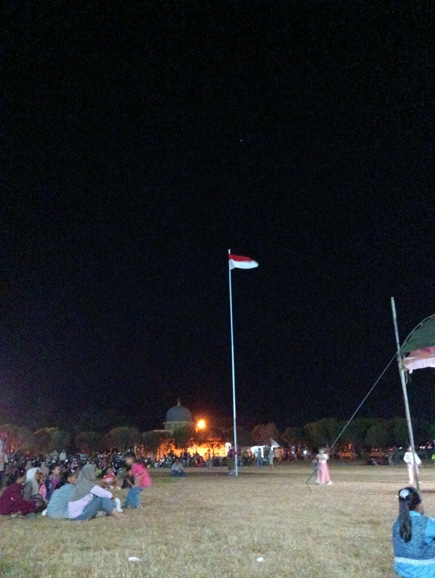The image depicts an outdoor gathering at night in Pematang Jaya, Indonesia. The scene features a crowd of people, some sitting on the grass and others standing, with a building that appears to be a mosque or similar structure in the background, illuminated by orange lights. A flag, likely the Indonesian flag, is prominently displayed on a flagpole. Several trees are scattered throughout the landscape. The overall atmosphere suggests a public event or celebration.