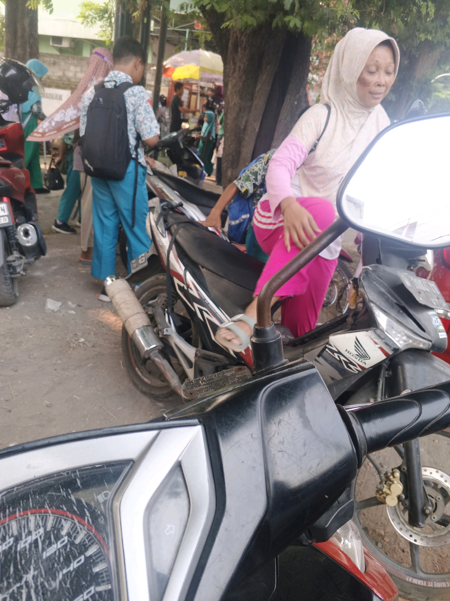 The image depicts a street scene in Krian, Indonesia. In the foreground, the handlebars and instrument cluster of a parked motorcycle are visible. To the left, another motorcycle with a silver exhaust pipe is parked. In the mid-ground, several individuals are present. A young male wearing a blue shirt, patterned with a light blue and white design, and turquoise pants, carries a black backpack. Next to him, a woman wearing a light-colored hijab and a light pink long-sleeved shirt over a bright pink outfit is seated on a motorcycle. Her left hand rests on her thigh. Further back, other people are visible, including a woman in a light blue hijab and green pants, and a person with a patterned backpack. The environment appears to be outdoors, with trees and buildings in the background. The ground is unpaved.