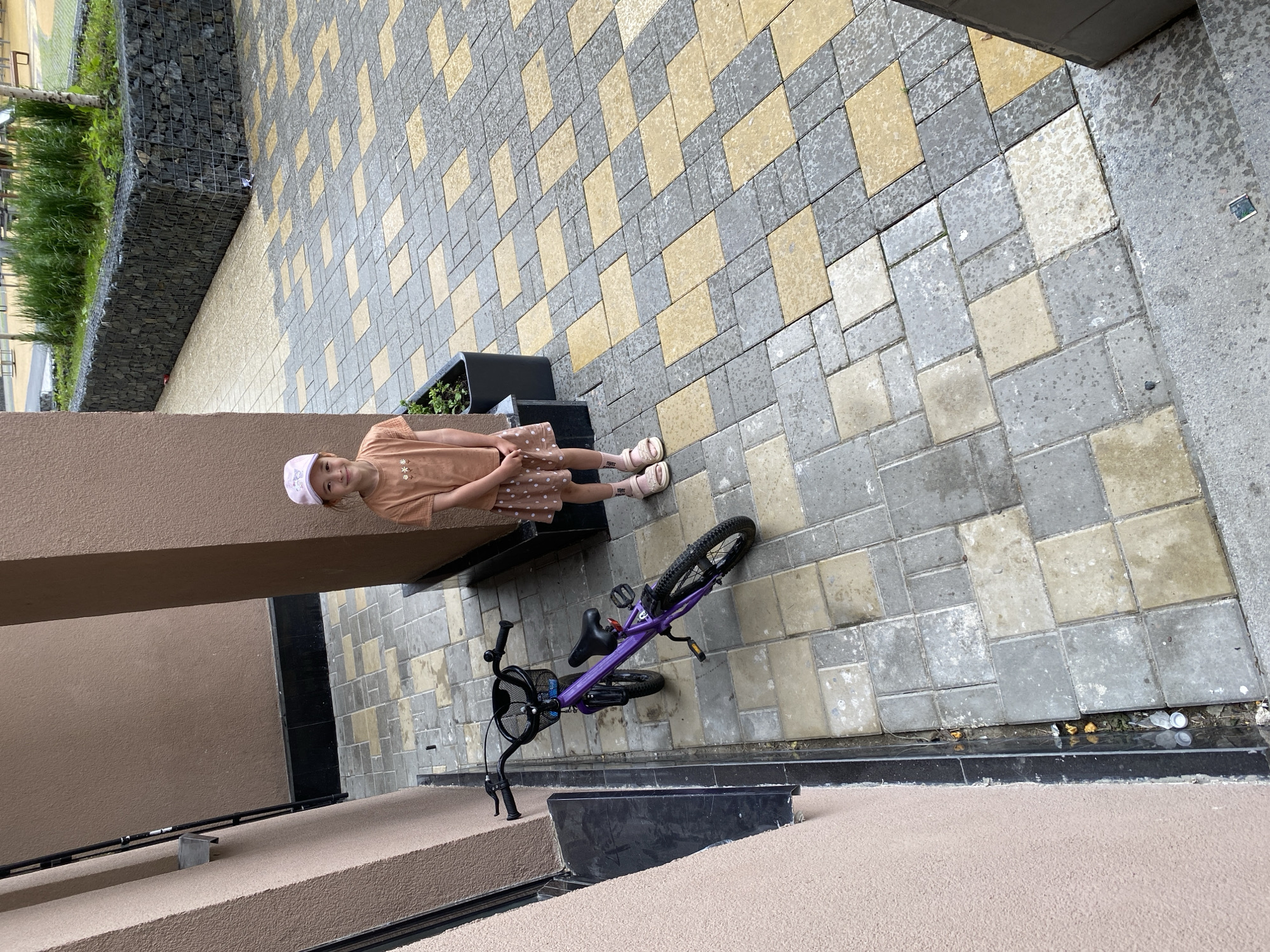 A young girl with a pink cap and an orange t-shirt stands next to a purple bicycle. She is wearing a polka-dot skirt and pink sandals. The bicycle is a children's model with a basket on the handlebars. They are standing on a patio paved with light gray and yellow rectangular tiles. To the left of the girl is a beige-colored building wall. Behind the bicycle, a dark planter box holds green plants. In the background, a gabion wall filled with stones surrounds a patch of lush green grass and trees. The scene appears to be in an urban outdoor setting, possibly a courtyard or public space in Yekaterinburg, Russia.