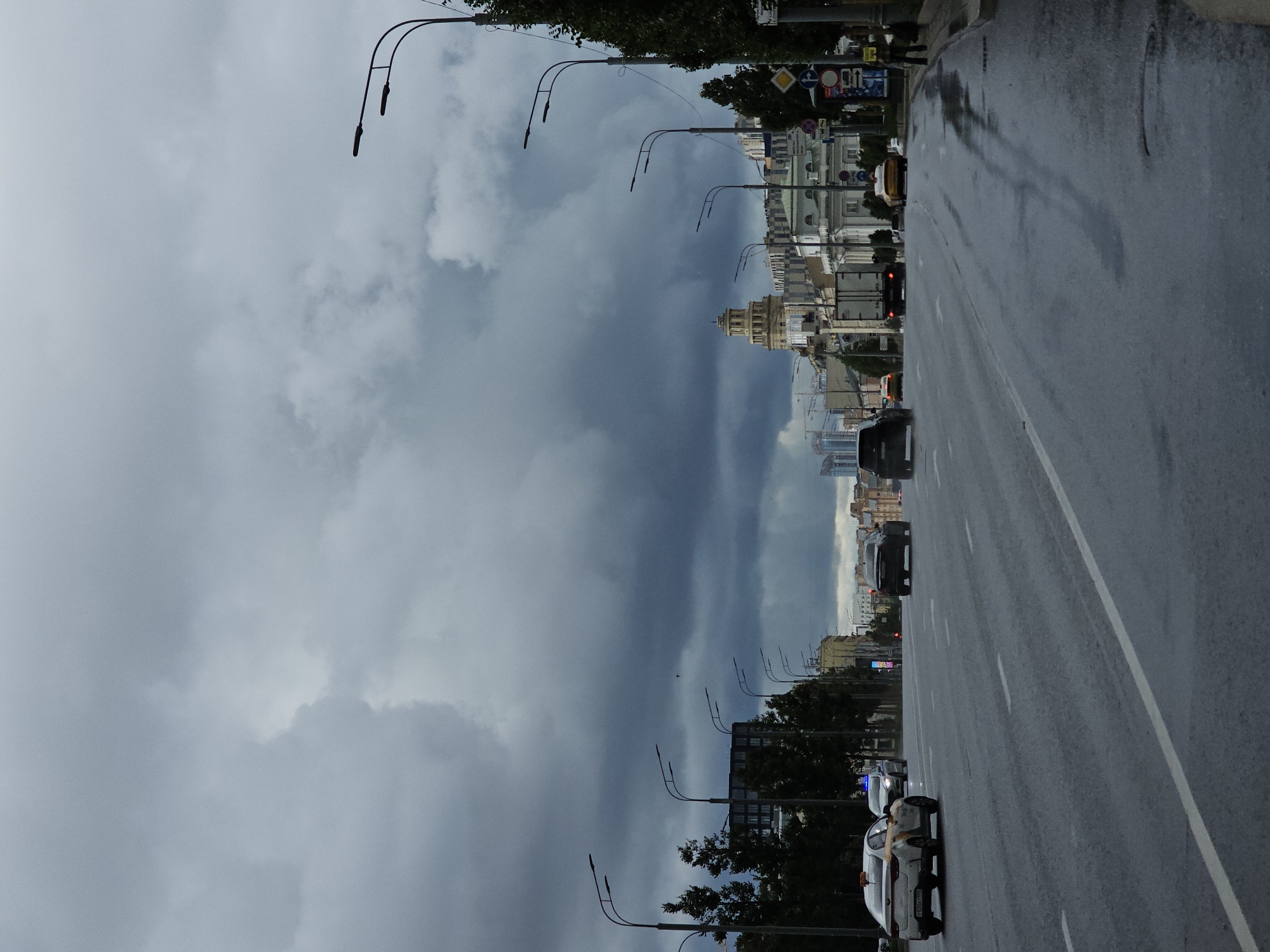 The image depicts a wide, multi-lane road in Moscow, Russia, under a heavy, overcast sky with dark, dramatic clouds that suggest impending or recent rain. The road surface is wet, with visible puddles and reflections, indicating damp conditions.
Several vehicles are present on the road. In the foreground, on the left lane, is a white and yellow taxi with the license plate "P720HY". To its right, further down the road, a dark-colored sedan is visible. Several other cars and a large white truck are also seen in the middle ground, traveling in the same direction. On the far right side of the road, a person is walking on the sidewalk next to a streetlamp.
Along the sides of the road, there are rows of streetlights, trees, and various buildings. To the left, a modern building with multiple windows can be seen. In the center of the image, a distinctive building with a tall, multi-tiered tower is prominent. Further in the background, other buildings of varying architectural styles, including some with classical elements, are visible. Several traffic signs are also present on the right side of the road.
The overall atmosphere is somber and slightly melancholic, largely due to the dark, cloudy sky and the wet road. The scene appears to capture a typical urban environment in Moscow during a period of inclement weather.