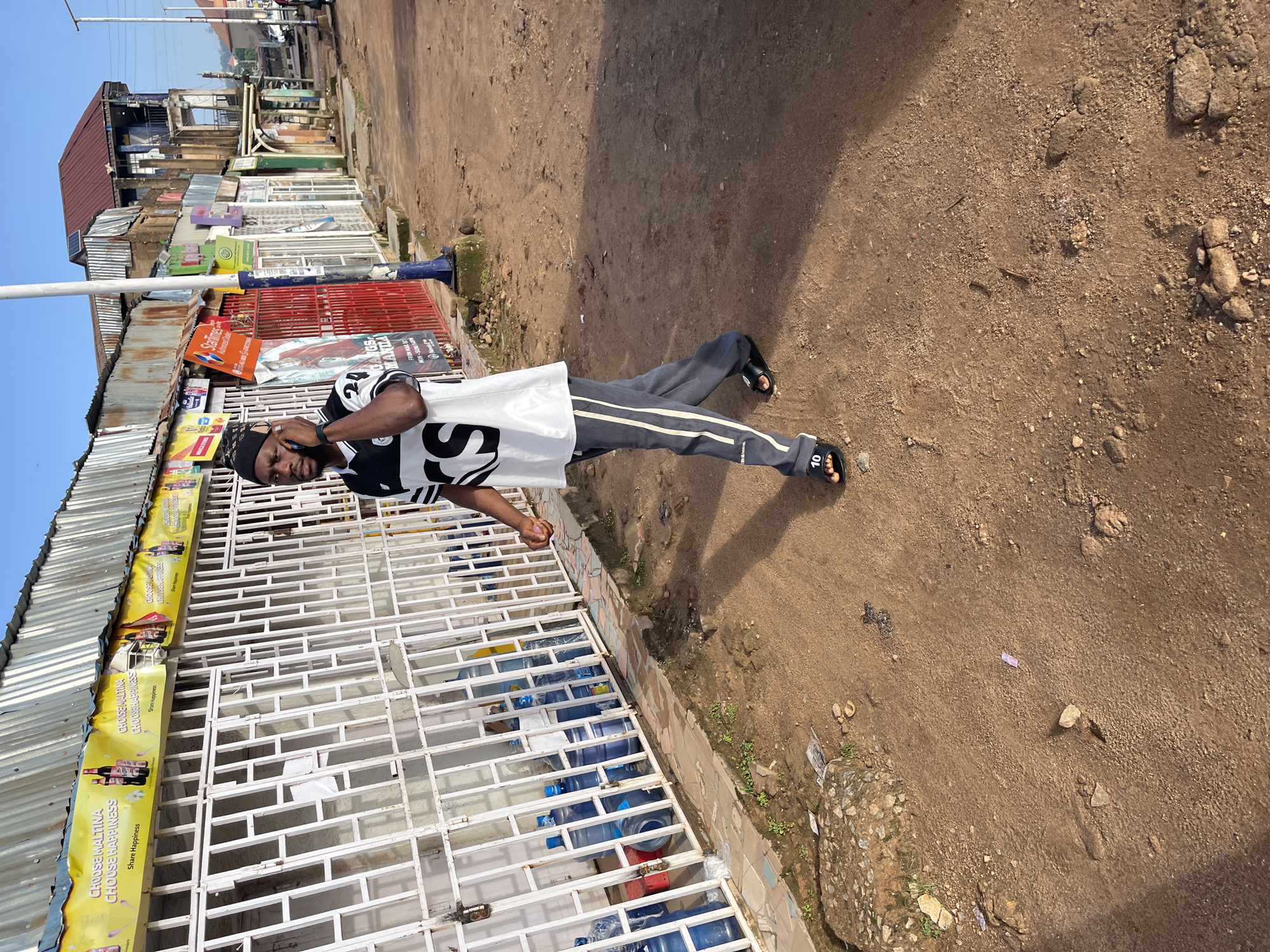A Black man wearing a black and white jersey and dark grey pants with white stripes is walking on a dirt road in front of a row of shops. He is holding a black mobile phone to his right ear and appears to be in a conversation. The shops have corrugated metal roofs and barred windows, with several large blue water containers visible behind the bars of one shop. Banners with the text "CHOOSE MALTINA CHOOSE HAPPINESS" are displayed above the shops. The road is dusty and uneven, with scattered debris. The sky is blue and clear. The scene appears to be in Gura Top, Nigeria.