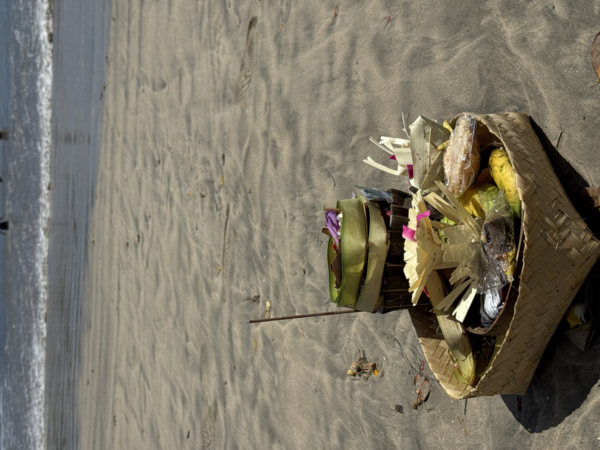 On a beach in Kuta Selatan, Indonesia, a Balinese Hindu offering rests on the sand. The offering is composed of a woven basket filled with fruit, flowers, and decorative palm leaves. An incense stick lies nearby. The tide is coming in along the beach, with waves visible in the background.