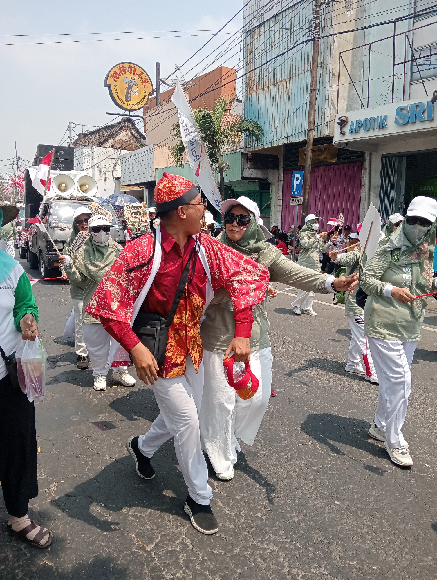 In Demak, Indonesia, a group of people participates in a street parade under a clear sky. The central figures are two individuals in the foreground, one wearing a vibrant red shirt and an elaborate red and gold patterned jacket, with a black crossbody bag. This person is moving dynamically, with their right arm extended and their body turned to face a woman to their left. The woman is dressed in a light olive green hijab, sunglasses, and white pants, and she is holding a red object with a plastic bottle attached. They appear to be interacting closely, possibly dancing or moving together as part of the parade.

Behind them, other participants are also dressed in similar light olive green long-sleeved shirts and white pants. Some are wearing white caps and sunglasses, and many are holding small Indonesian flags or other parade props. A truck with speakers mounted on top is visible in the background, suggesting a sound system is part of the procession.

The parade takes place on a paved street, with buildings lining the right side of the road. One building displays a prominent sign for "APOTIK SRIWIJAYA" and another for "MR. D.I.Y. Always," a hardware store chain. The environment suggests a commercial or residential street in a bustling urban area. The overall scene depicts a lively community event.