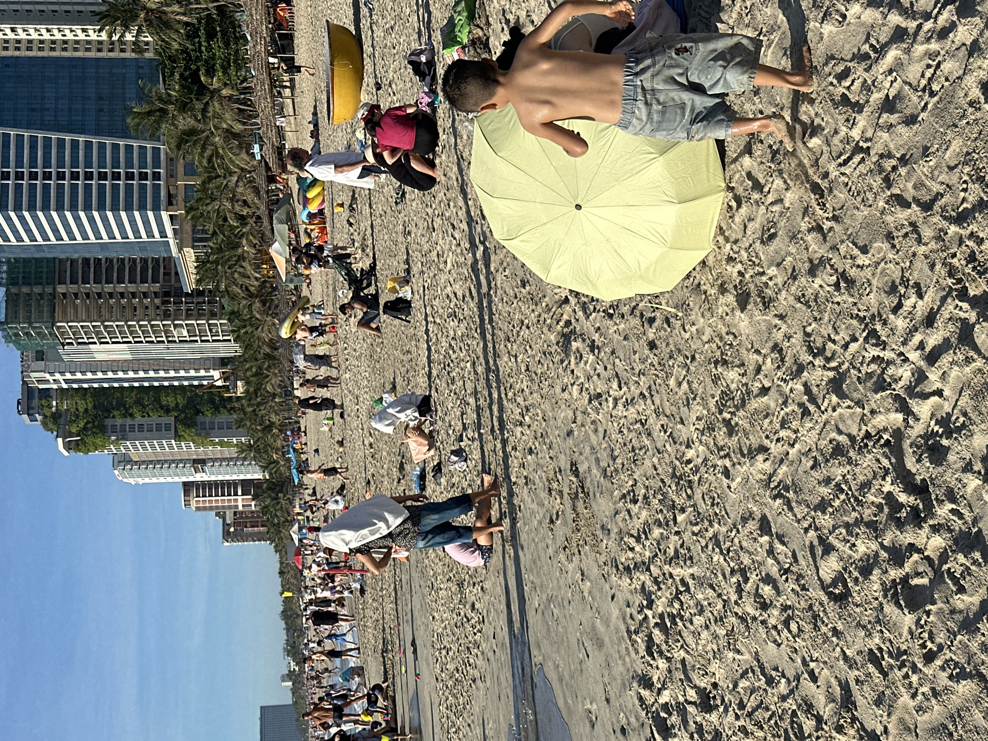 The image captures a bustling scene on a sandy beach in Da Nang, Vietnam, with modern high-rise buildings lining the background. The beach is populated with numerous individuals engaged in various activities, suggesting a warm, sunny day.
In the foreground, a young boy with light brown skin, wearing denim shorts, is running barefoot on the sand. He is facing away from the camera and towards a light green umbrella that is open and lying on the beach. His body is turned slightly to his left, and his arms are extended backward, indicating motion. His skin appears wet and glistening.
Behind the boy, the sand is textured with footprints and small ripples, indicating recent activity and perhaps the receding tide. Further back on the beach, various groups of people are scattered. Some are sitting, others are standing, and some appear to be interacting with each other. A woman wearing a white head covering and a patterned shirt is standing with her hands on her hips, looking towards the left. A person wearing denim shorts and a blue t-shirt is standing in the water at the edge of the beach.
A yellow boat-shaped object, possibly a basket boat, is visible on the sand. Several beach umbrellas and loungers are also present, along with various bags and personal belongings. The overall environment suggests a popular tourist destination, with the clear blue sky and palm trees contributing to a tropical atmosphere. The tall buildings in the background, some under construction and others completed with glass facades, denote the urban development of Da Nang.