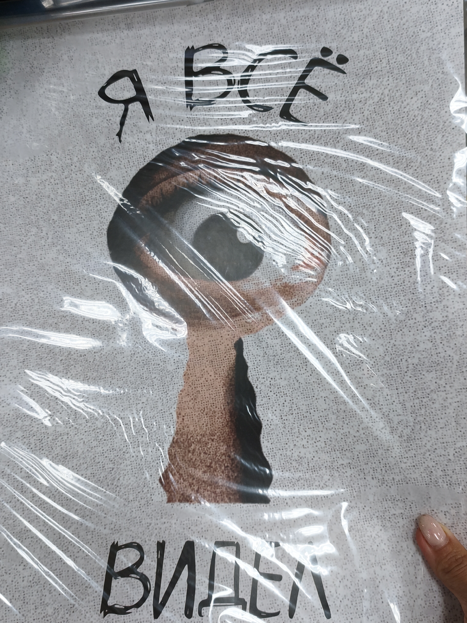 The media file shows a close-up of a poster or print, partially covered by a protective plastic film. The artwork features bold, stylized Cyrillic text at the top and bottom. The top text reads "Я всё" and the bottom text reads "виден". In the center of the image is a graphic illustration of a large eye, positioned within what appears to be a keyhole shape. The eye is rendered with brown irises and a dark pupil, looking directly at the viewer. The keyhole shape is formed from a reddish-brown, textured material. The background of the artwork is a light-colored, textured surface, possibly representing paper or fabric. The plastic film creates reflections and distortions, making the text and image slightly less clear. A person's hand with a light pink nail polish is visible in the lower right corner, touching the plastic film, providing a sense of scale and indicating the object is being handled. The overall impression is that of a piece of graphic art with a somewhat surreal or observational theme. There are no explicit cues about the time of day, weather, or a specific indoor or outdoor setting beyond it being an environment where such an object might be displayed or stored.
