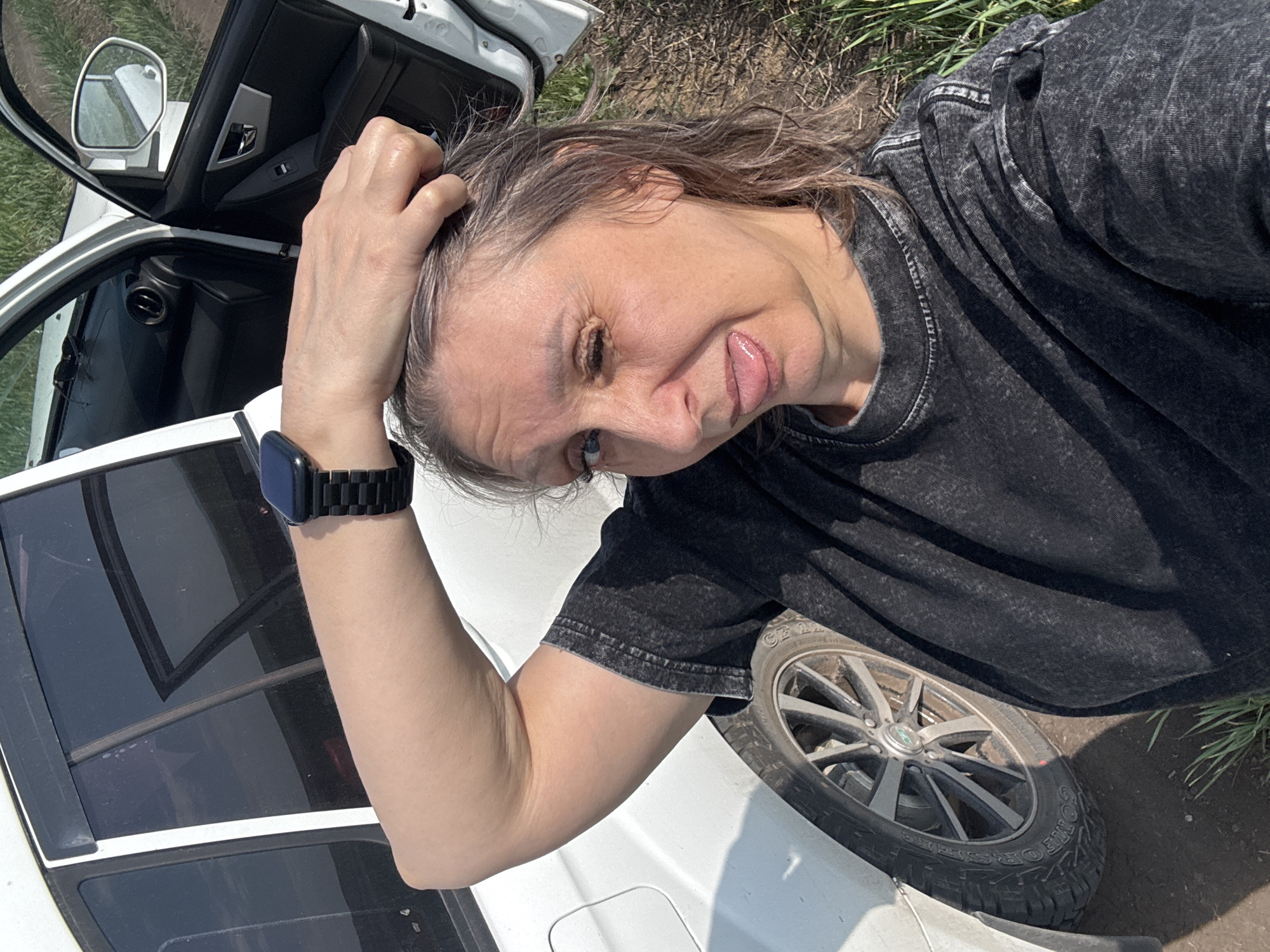 A woman is taking a selfie outdoors in the village of Troitskoye, Russia. She is standing next to an open white car, with the front passenger door ajar. The woman is wearing a dark grey t-shirt and has her left arm raised, resting her hand on the top of her head. She is winking with her right eye and has her tongue sticking out. The car's interior, including the black dashboard and part of the passenger seat, is visible. The background shows a grassy area and a dirt road. The image is taken on a sunny day.