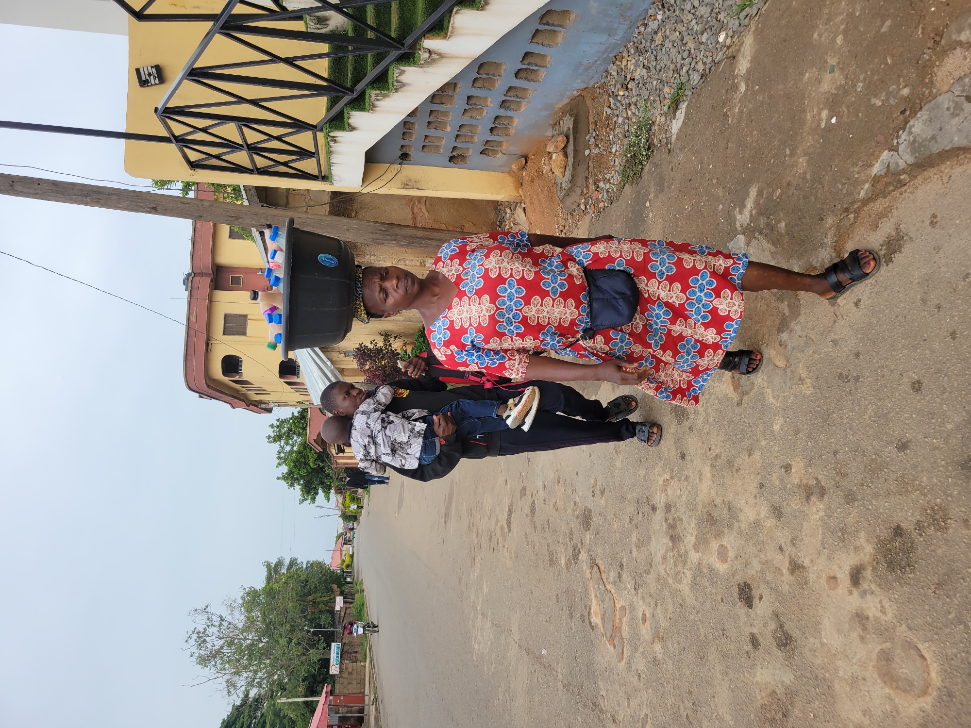 An outdoor shot on a paved street in Jos, Nigeria, depicts a woman in a red floral dress, carrying a large black basin full of bottled beverages on her head. A man, closely following her, holds a young child. The backdrop includes buildings, some with distinctive architectural features, alongside trees lining the street. A sign with the word "Restaurant" is visible. The scene suggests daily life and activity within the community.