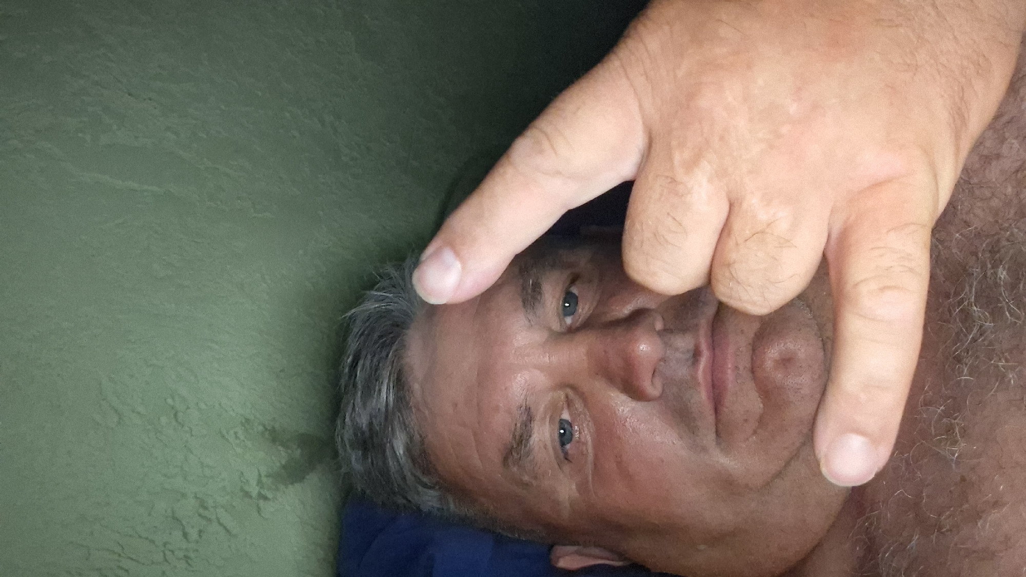 A close-up, selfie-style photograph shows a man with graying hair and blue eyes. He is looking directly at the camera. His right hand is raised, with the index finger and pinky extended in a gesture commonly associated with rock and roll. The background is a textured, green wall. The image appears to have been taken indoors. There is no information within the image to indicate the location is Voinova, Moldova.