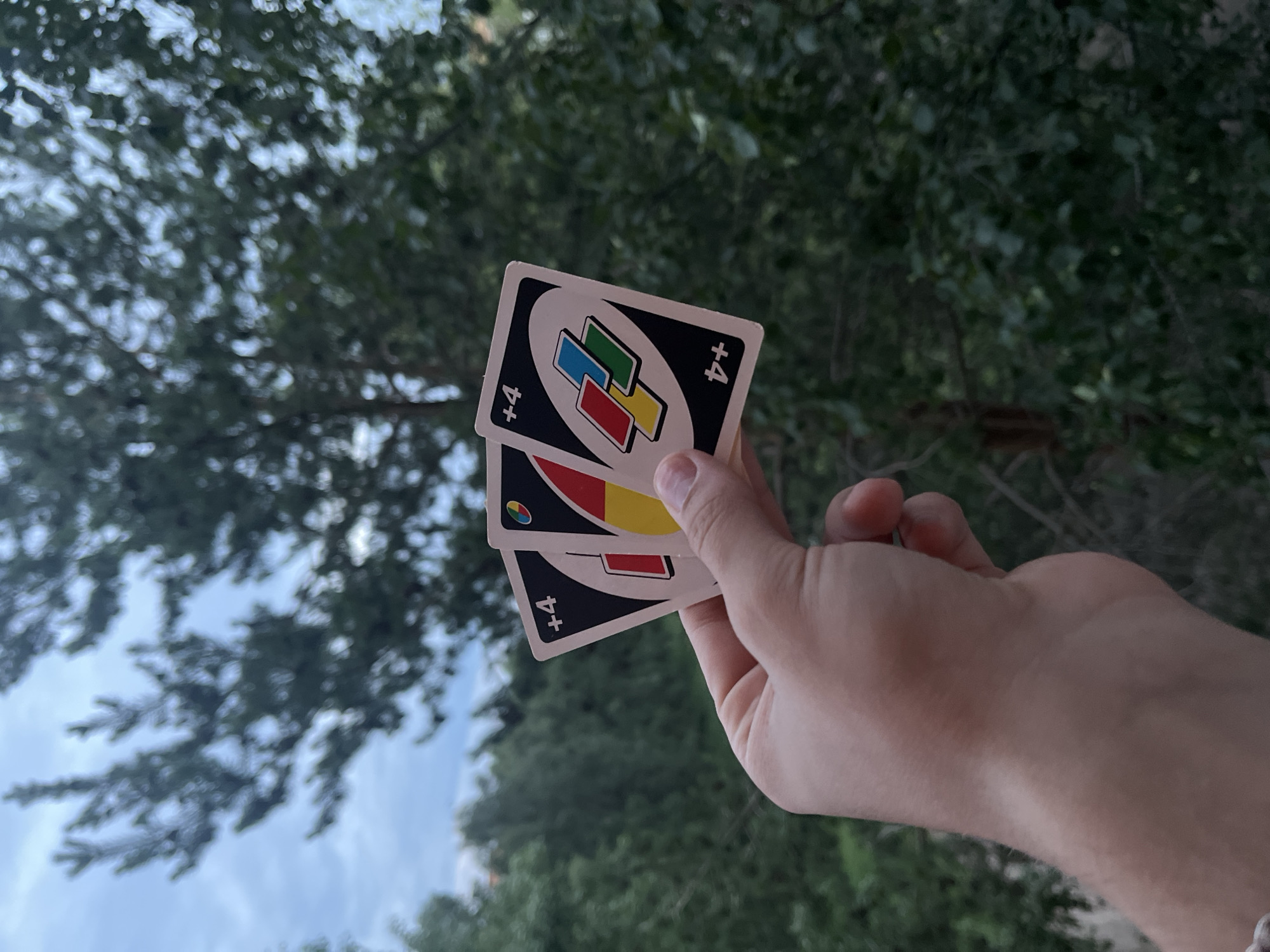 A person's hand holds three Uno Wild Draw 4 cards outdoors. The cards are fanned out, with the top card displaying the text "+4" in the upper left and a red rectangle on a black background in the center. The second card, partially obscured, also shows "+4" and a portion of a red and yellow circular design with a blue segment. The third card, most visible, features "+4" in both upper and lower right corners, with a stylized circular motif containing overlapping red, blue, yellow, and green squares on a black background. The background consists of blurred green foliage and a cloudy sky.