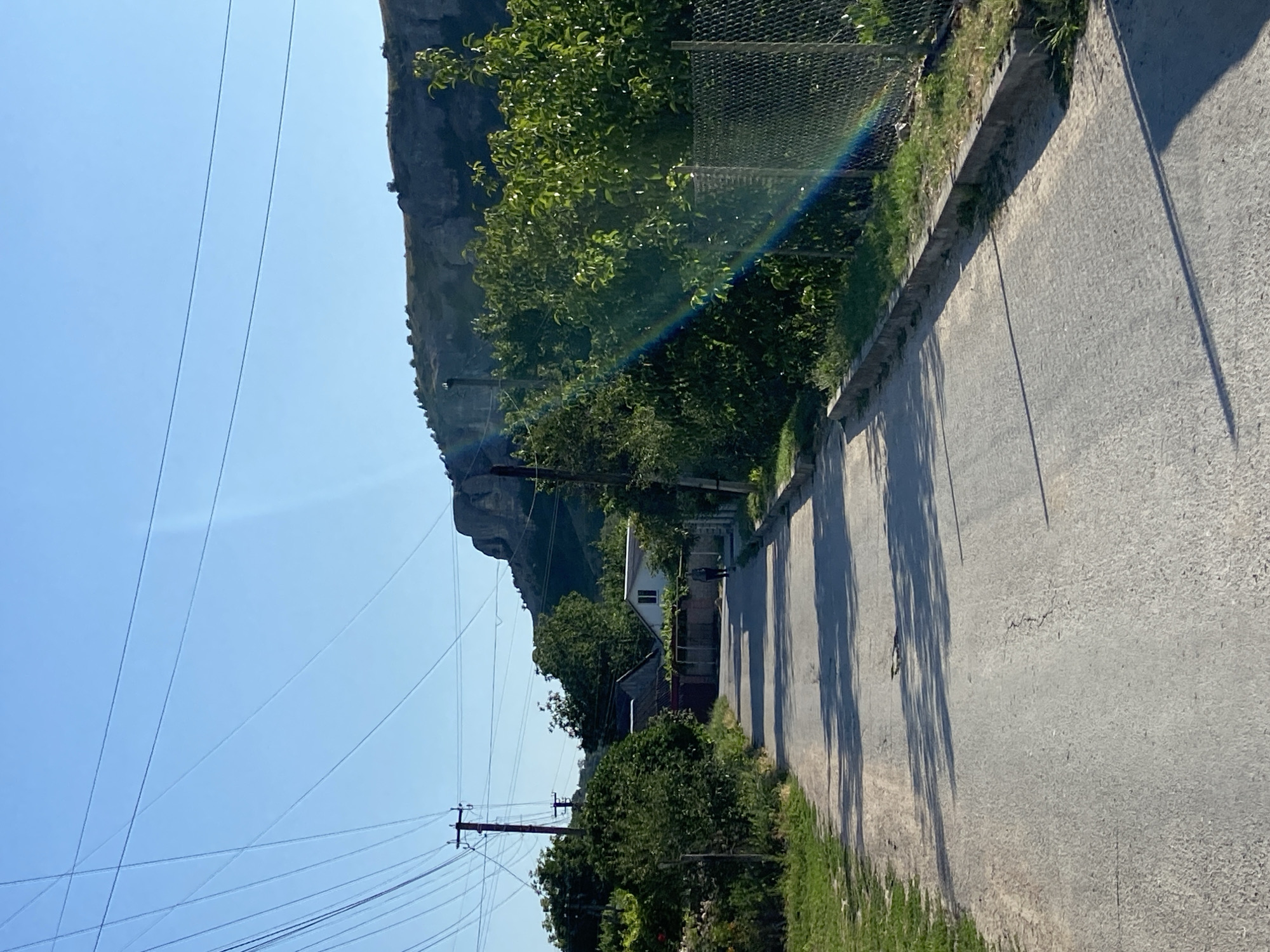 The image depicts a rural road in the Krasnomakskoye rural settlement, Ukraine. The road is paved with asphalt and shows signs of wear, with cracks and patches visible. Lush green trees and bushes line both sides of the road, with a chain-link fence running along the right side. In the distance, a person is walking away from the camera down the road. To the left of the road, there are some houses with red roofs and one house with a white facade. A prominent cliff face looms in the background, under a clear blue sky with some overhead power lines crisscrossing the frame. A faint rainbow is visible on the right side of the image, appearing as a multicolored arc in the sunlight.
