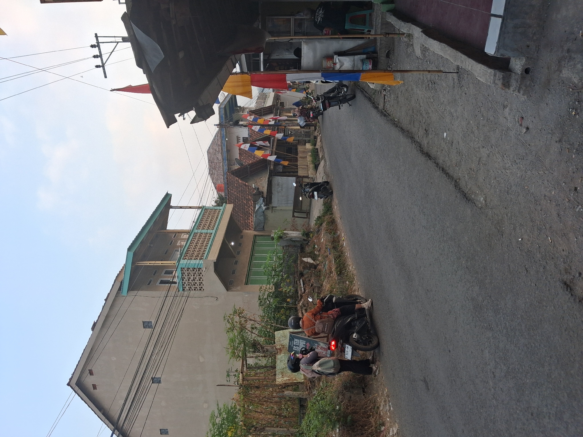 The image depicts a street scene in Bantarkawung, Indonesia, likely in a residential area or a small commercial strip. The road is paved and appears to be a two-lane street. On the left side of the road, there is a light-colored, two-story building with a balcony that has teal railings. Electrical wires are strung across the facade of the building. In front of this building, there is a motorcycle with two people wearing helmets. One person is seated on the motorcycle, and another is standing next to it. A sign with text is visible behind the standing person. There are also various plants and some debris on the side of the road.
On the right side of the street, there are several buildings with tiled roofs. Many colorful flags, primarily red, white, and yellow, are strung across the street and attached to poles, suggesting a festive occasion or a local event. One building on the right appears to be a shop with an open front. Inside, a person is seated on a stool, possibly attending to customers or engaged in an activity. Various containers, including a large white bucket labeled "GIVI", are visible near the shop entrance. The ground on the right side of the street is uneven and appears to be made of dirt and gravel, with some concrete structures. The sky is partly cloudy, suggesting it is either early morning or late afternoon.