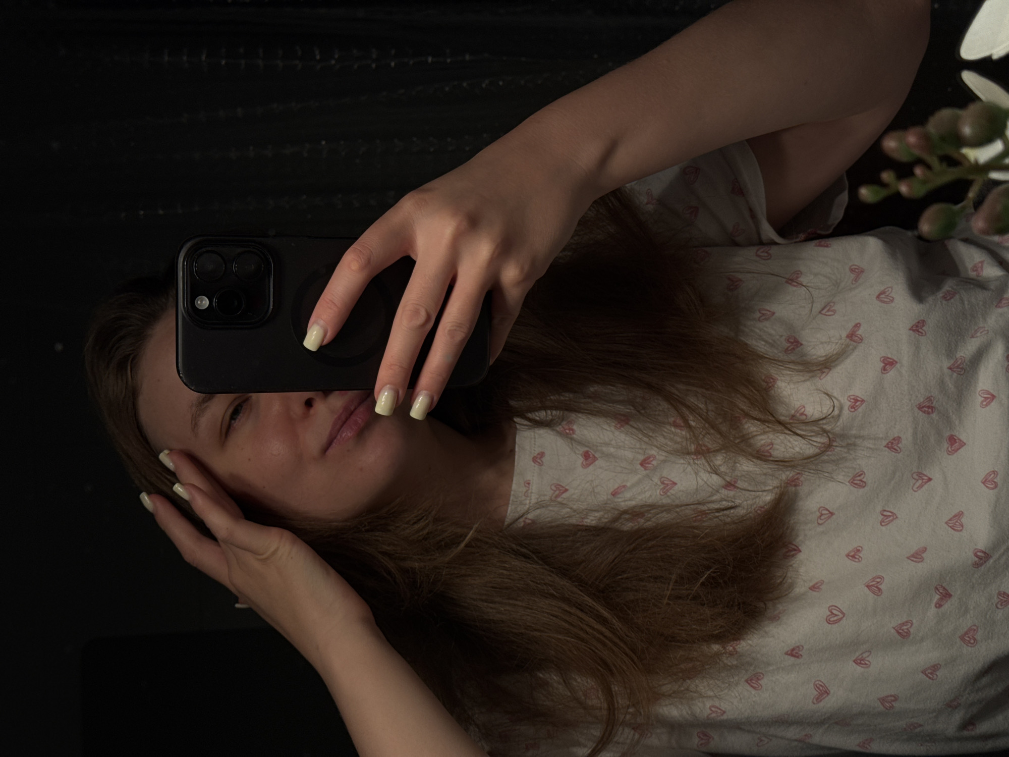 A person is visible in a reflective surface while holding a black smartphone. The person has long, light-brown hair. Part of their face is visible; the rest is obscured by the phone. The visible skin tones are fair. One hand holds the phone, with pale yellow manicured nails visible. The other hand is partially visible near the face. The person is wearing a light-colored garment printed with red hearts. Reflected in the background is a dark surface, possibly a mirror or glass. To the right, there is part of a plant with pale green seed pods, possibly artificial. The setting does not appear to be in Vladimir, Russia.