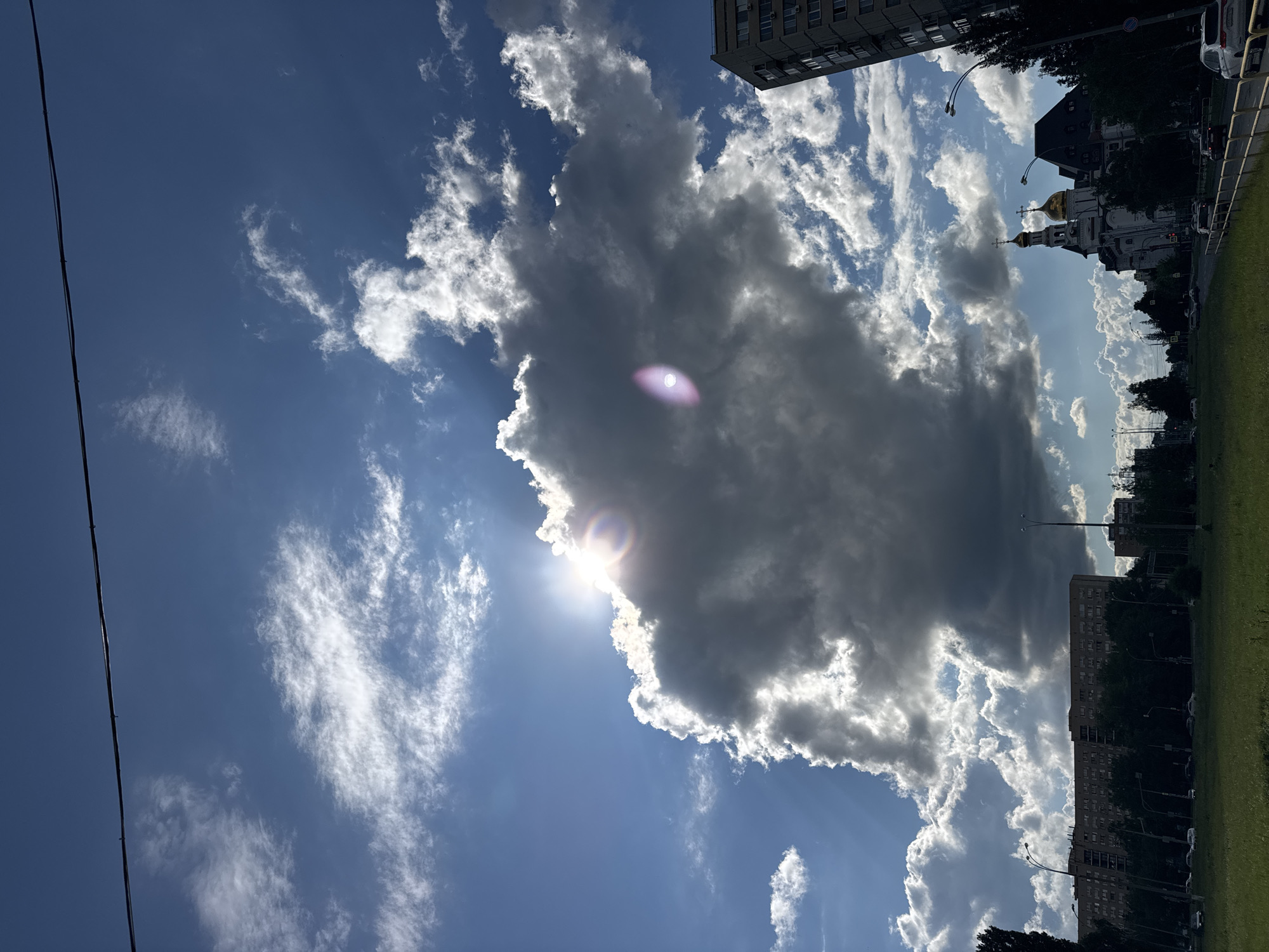 The image captures a dramatic sky over an urban landscape. A large, dark cloud dominates the center, partially obscuring a bright sun that creates a lens flare effect. Rays of sunlight pierce through the edges of the cloud, illuminating lighter clouds around it. In the lower portion of the image, a street with moving cars is visible, bordered by trees and a grassy area. To the right, a modern multi-story building stands tall, and further down the street, a building with golden domes, likely a church, is present. The scene appears to be set in the afternoon under a dynamic sky.