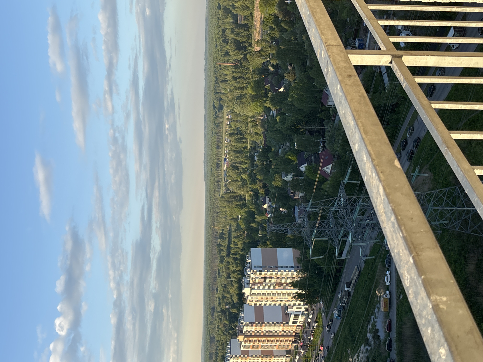 The image shows a view from a high vantage point, possibly a tower or tall building, overlooking a residential area and forest in Vsevolozhsk, Russia. The sky is blue with scattered clouds. Below, a dense forest stretches towards the horizon. Closer to the viewer, there are residential buildings, including multi-story apartment complexes. Roads with cars are visible, along with power lines and support structures. A metal railing, likely part of the structure from which the photo was taken, dominates the foreground. There are no people distinctly visible, though possible human presence is suggested by the cars and buildings. The environment is urban, with residential areas surrounded by natural forest.