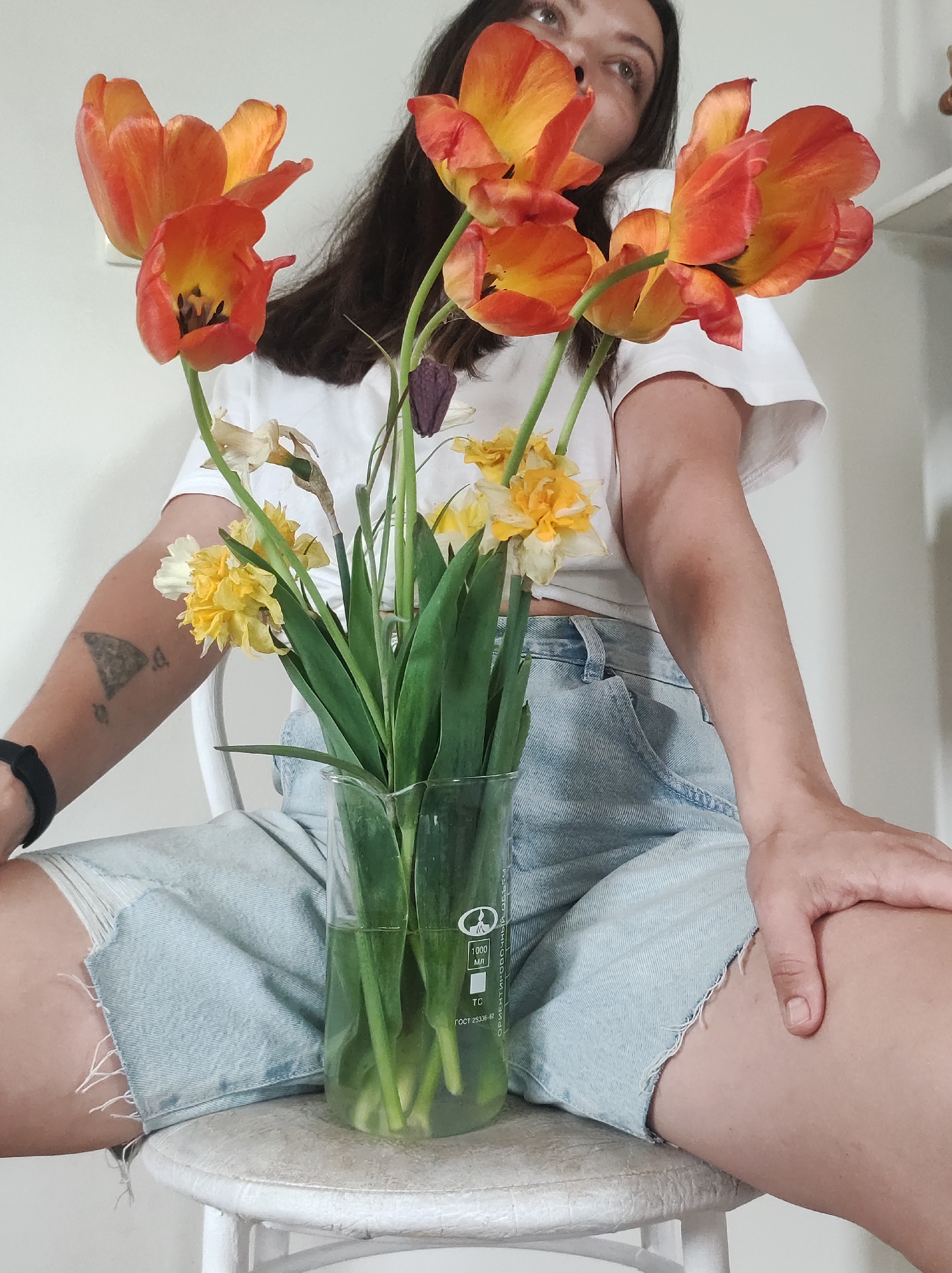 A person sits on a white chair, their legs crossed. They wear a white t-shirt and light blue denim shorts with frayed edges. A clear glass vase filled with water stands between their legs, holding an arrangement of orange and yellow tulips and yellow daffodils. The vase is marked with "1000 ml" and other Cyrillic text. The person's right hand rests on their right leg. Their left wrist has a black band, and there is a tattoo on their left forearm. The background is a plain white wall. There is no indication of the location being in Фоки, Russia.
