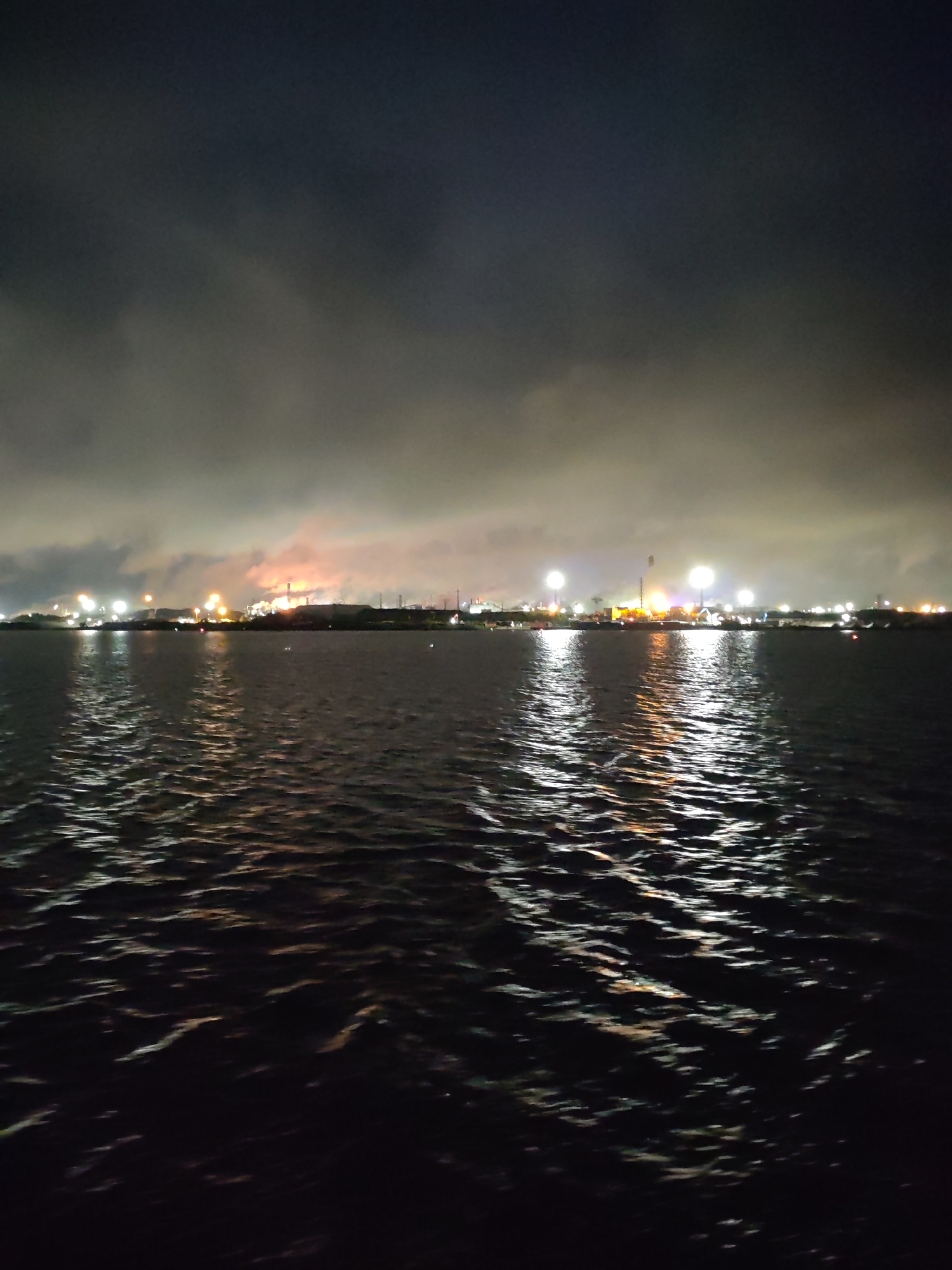 The image depicts a nighttime view of the Sheksna River in Cherepovets, Russia. The foreground is dominated by the dark, rippling water of the river. In the background, the city skyline is visible, illuminated by numerous lights. Some lights appear brighter and more concentrated, suggesting industrial or commercial areas, while others are more dispersed, likely representing residential zones. The sky is overcast, with clouds obscuring the upper portion of the image. The overall atmosphere is dim and subdued, accentuated by the dark tones of the water and the muted glow of the city lights. There are no people visible in the frame.
