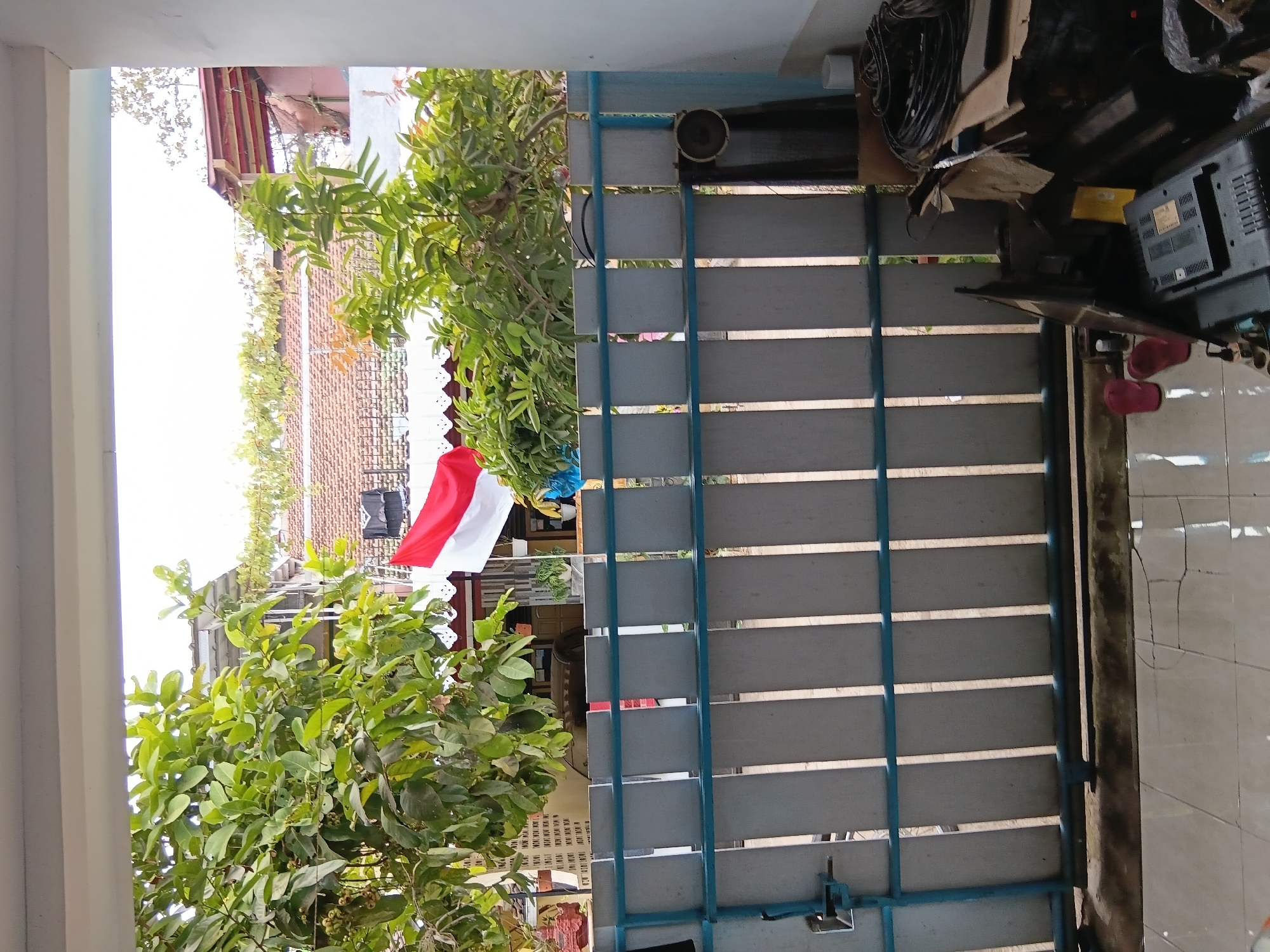 The image depicts a view through a blue and gray fence, looking towards a residential area in Tanggul Wetan, Indonesia. A red and white Indonesian flag is prominently displayed, attached to a building visible behind the fence. Lush green trees with visible fruit or small buds are to the left and right of the flag, obscuring parts of the buildings. The fence itself is made of vertical gray planks with horizontal blue bars. To the right of the fence, in the foreground, is a collection of discarded electronics, including a television set and coiled cables. A pair of red flip-flops are visible on the tiled floor in front of the fence, to the left of the electronic debris. The overall scene suggests an outdoor residential setting with a mix of nature and discarded household items.