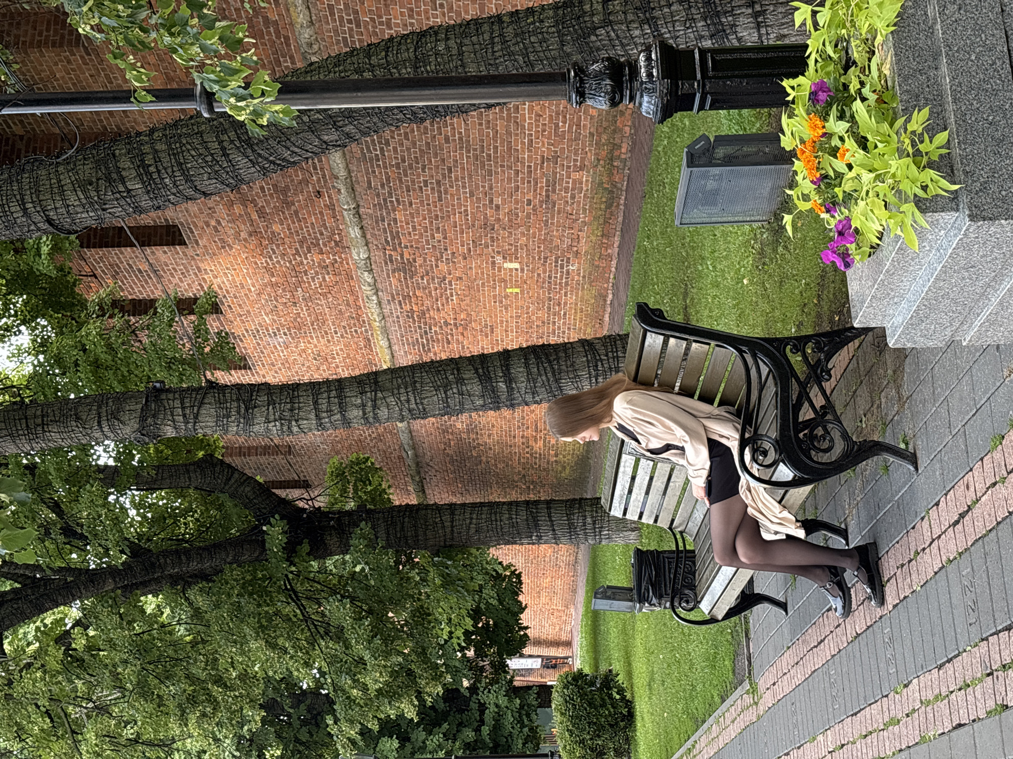 A young woman with blonde hair is sitting on a park bench in an outdoor setting. She is wearing a light-colored trench coat over a dark outfit, black tights, and dark shoes. Her head is bowed, and she appears to be looking down. The bench is made of wood and black metal.
Behind the bench, there is a granite structure with a planter containing vibrant orange and purple flowers. A black lamppost is also visible. The ground is paved with a pattern of grey and pinkish-brown bricks, and to the left of the path, there is a grassy area. In the background, large trees with dark bark wrapped in netting are prominent, and behind them is a brick wall. The scene suggests a park or a public garden. The lighting indicates it is daytime, likely overcast or shaded due to the lack of strong shadows.
No visible text is present in the image. The overall mood seems quiet and contemplative.