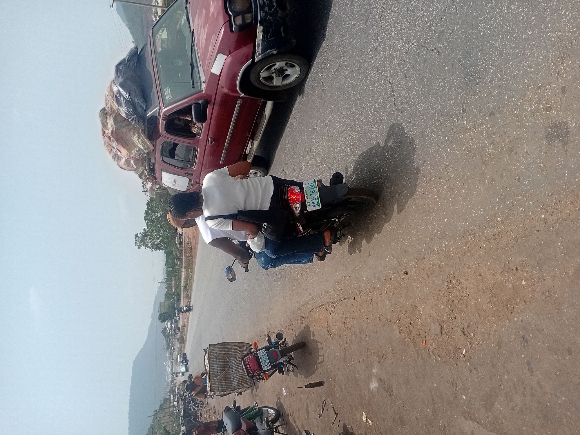 The image captures a vibrant roadside scene in Lokoja, Nigeria, under bright daylight. The perspective is slightly elevated, looking down at a diagonal stretch of an asphalt road.
In the immediate foreground, two individuals are riding a black motorcycle. The person in front wears a light-colored top, while the rider behind wears a white t-shirt and blue jeans, carrying a dark bag. The motorcycle's rear license plate clearly displays "KOGI APA-764QC".
Adjacent to the motorcycle, a red pickup truck is visible, its open bed heavily laden with numerous sacks and bundles, some covered in dark plastic. Partial text, "CLEP FOO...", can be deciphered on one of the sacks. A person's arm is faintly visible through the driver's side window. Both the truck and the motorcycle cast distinct, dark shadows on the sunlit asphalt, indicating a clear, sunny day, likely mid-morning or early afternoon.
Further down the road, to the right, another motorcycle with a large, striped basket-like carrier on its rear is parked, its license plate reading "KOGI 707XAAS". Scattered along the dirt shoulder are other motorcycles and people, suggesting a busy local hub or market area.
The background reveals a long, winding road flanked by sparse trees and distant structures. A prominent feature of the landscape is a hazy, multi-layered mountain range stretching across the horizon, characteristic of Lokoja's hilly terrain. The overall scene depicts daily life and transport activity in this region, with goods being moved by truck and people commuting on motorcycles.
