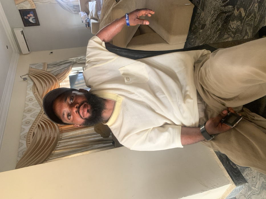 An adult male is seated indoors, likely within a residence in Eika-Adagu New Layout, Nigeria. He is positioned facing the camera, wearing a black baseball cap, a cream-colored V-neck short-sleeved top with a lighter yellow trim and a left chest pocket featuring a small metallic emblem, and light brown trousers. He has a full beard and is looking directly forward. A black wristwatch is present on his left wrist, and a blue silicone wristband with "Bungie" inscribed in white text is on his right wrist. His left hand holds a black feature phone. He is seated on a chair with a dark-colored back, positioned in front of a light cream-colored wall. The background features light brown/gold patterned curtains, an air conditioning unit mounted on the wall to the right, and a framed portrait below the AC unit. A beige upholstered sofa is partially visible behind him to the right. The floor is dark patterned marble or tile.