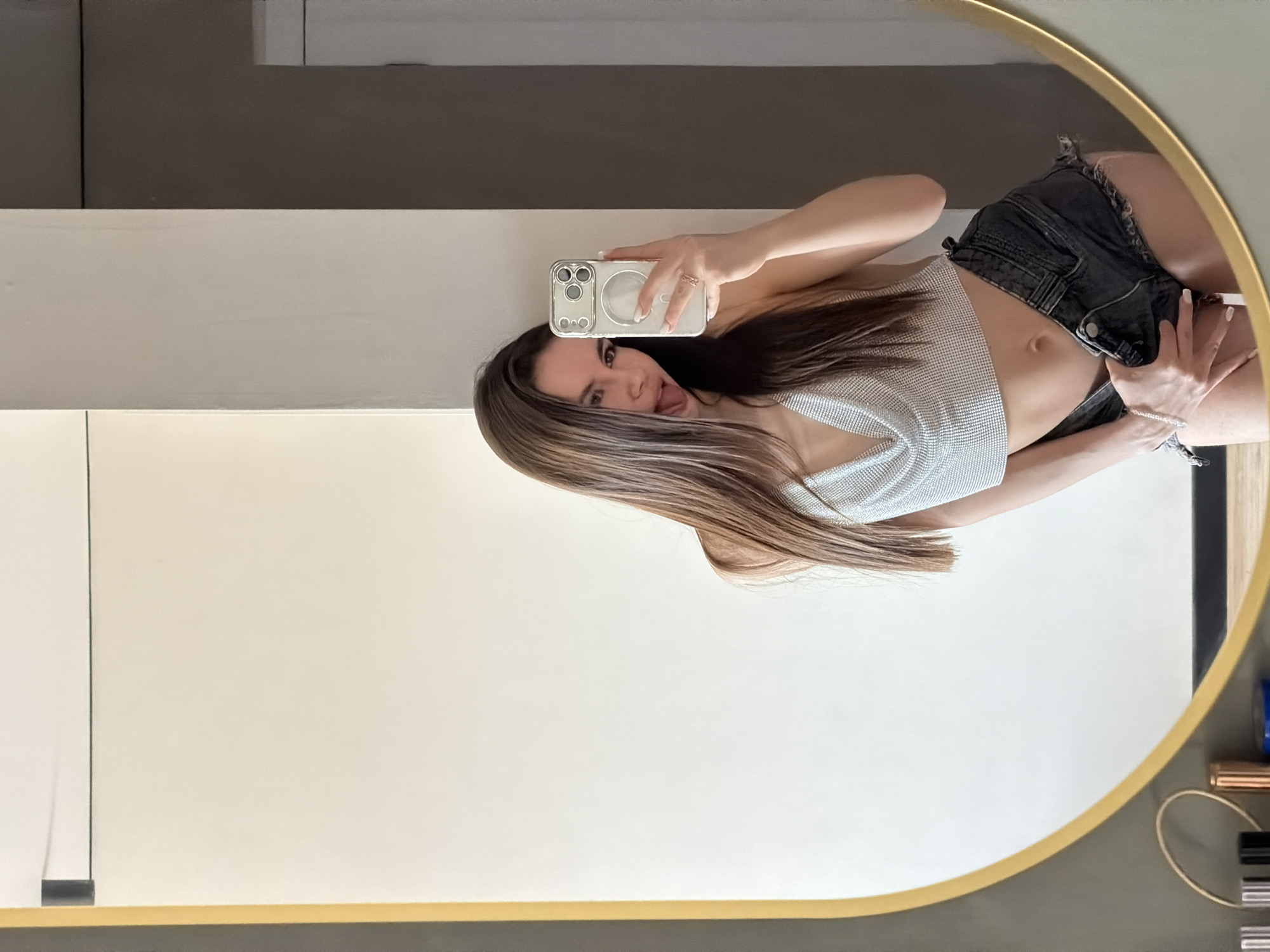 A woman is taking a selfie in a round mirror. She is wearing a silver top and black denim shorts. She is sticking her tongue out. The background appears to be a wall in an interior space. The location is unknown.