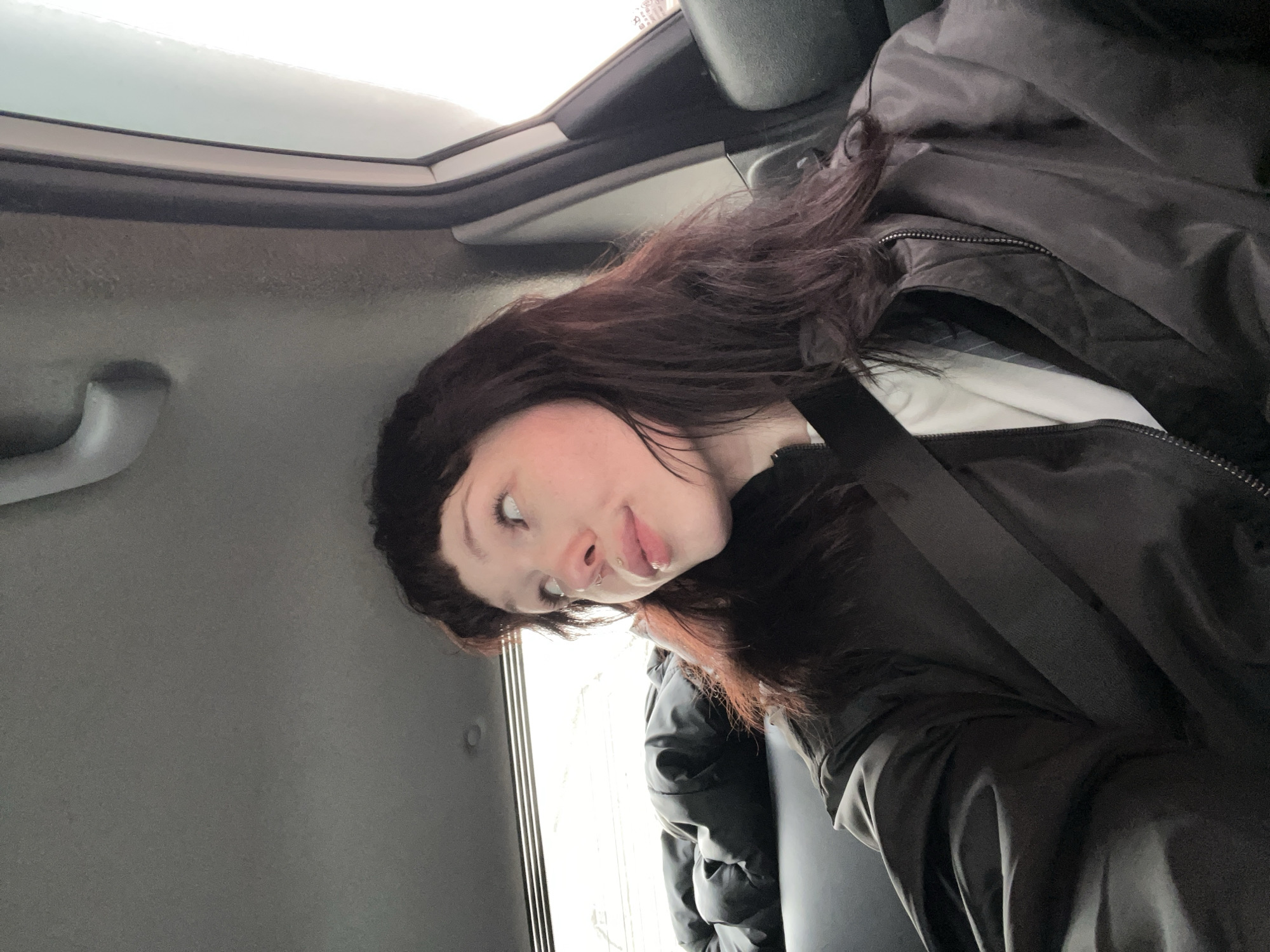 A person is seated in a car. They are wearing a dark jacket and a seatbelt is fastened across their chest. The car interior is grey. A window and part of the car's roof are visible. The person has dark hair with auburn highlights and facial piercings. The location is not identifiable as Kazan, Russia, from the provided image.