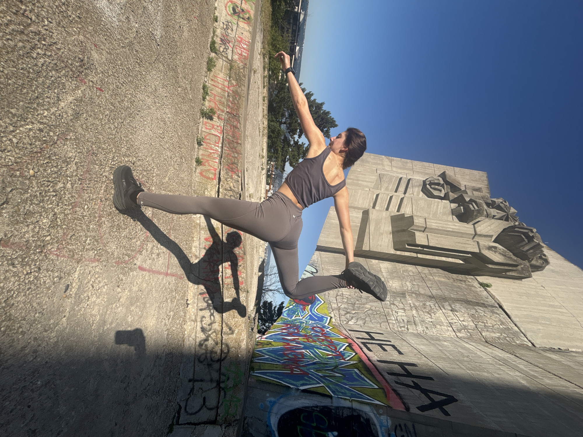 A young woman performs a yoga pose, dancer's pose (Natarajasana), on a concrete platform in front of a large Brutalist monument. The monument, with its imposing concrete structure and sculpted figures, is covered in graffiti. The woman, wearing a grey athletic set and black sneakers, is in mid-pose, with her right leg extended behind her and her left leg supporting her. Her arms are outstretched, with her right hand reaching towards her right foot, and her left arm extending forward. Her shadow is cast on the ground behind her, mirroring her pose. The background shows a clear blue sky and a distant cityscape with trees. The concrete surface beneath her is weathered and shows signs of age, with some graffiti and patches of dry grass. The overall scene suggests an outdoor urban setting, likely a public space with historical or artistic significance in Varna, Bulgaria.