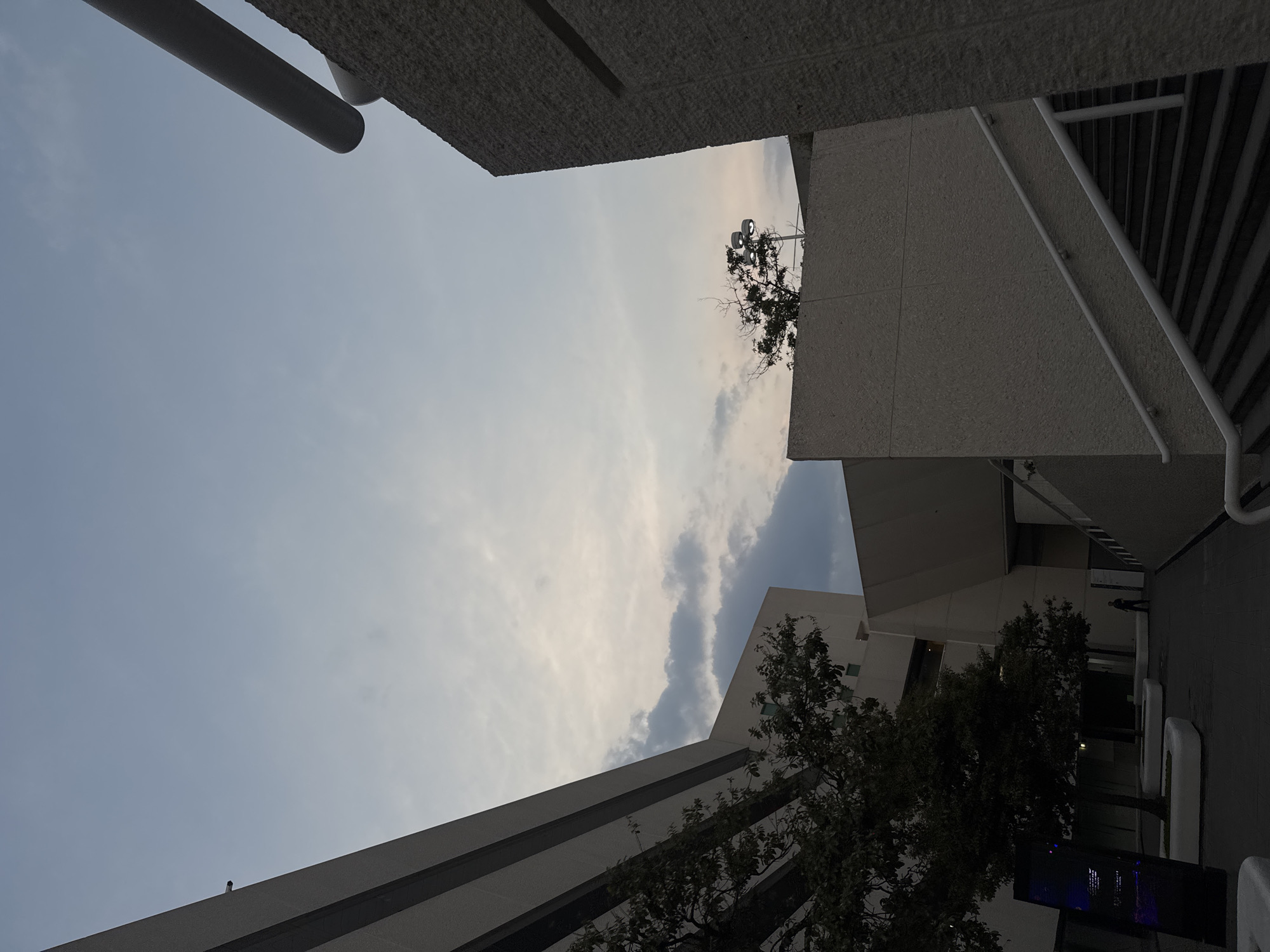 The image depicts a view upwards in an urban setting, likely in Heroica Puebla de Zaragoza, Mexico. The sky is visible, with a mix of blue and light, wispy clouds. Buildings frame the sky: one with a textured, concrete-like surface and rounded metal bars, one with striped patterns, and a tall, modern building with trees in front of it. A street light pole is visible.