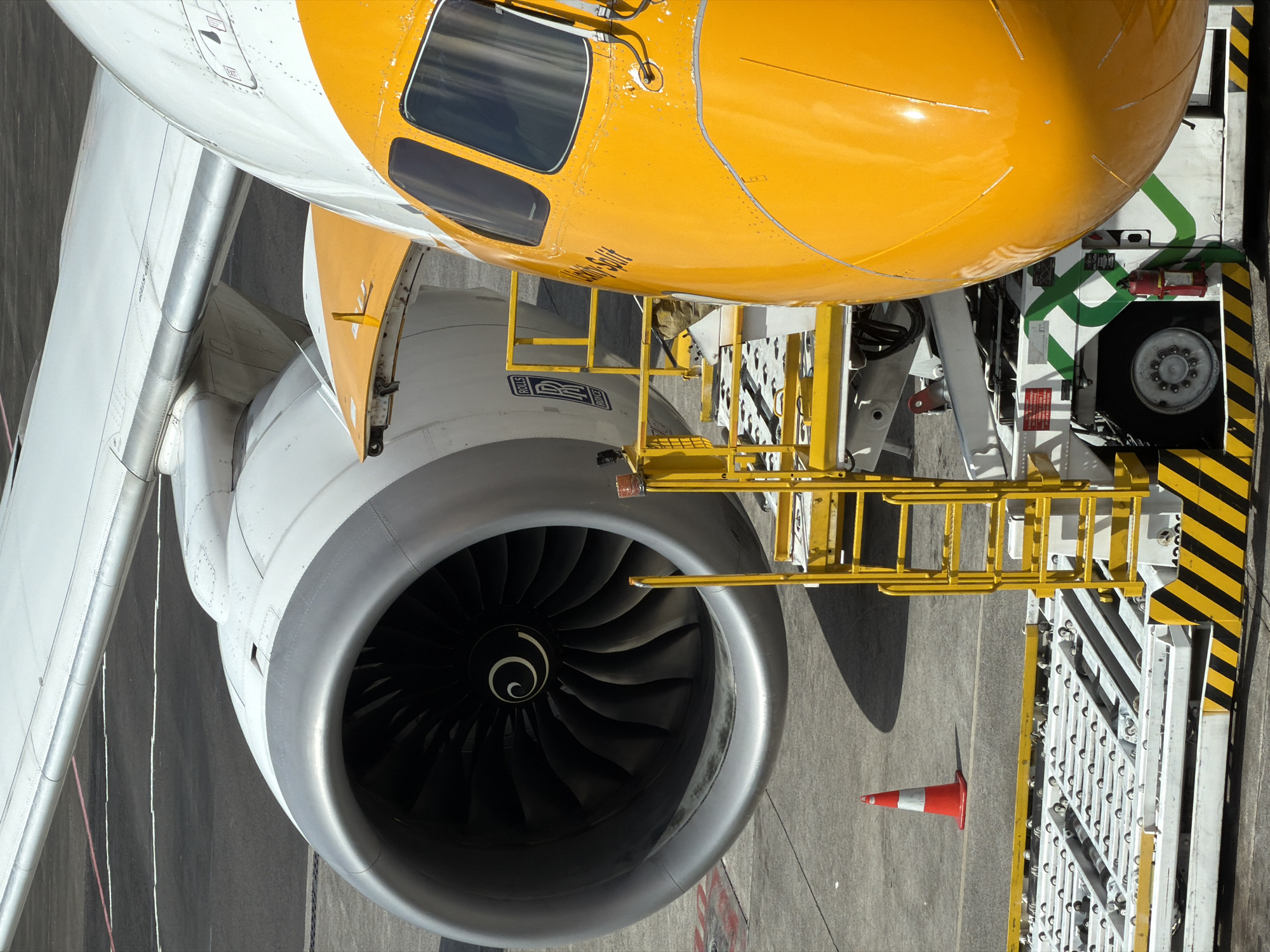 The image shows a close-up of an airplane's engine and nose on an airport tarmac. The engine is large and metallic, with visible fan blades inside. A portion of the airplane's white wing extends from the top left of the frame. The nose of the aircraft is bright yellow with the text "Skippy-Split" visible on it. A yellow boarding ladder is positioned next to the aircraft, leading up to an open hatchway on the engine nacelle. In the foreground, a loading platform with rollers is visible, along with a red and white traffic cone to the left. The tarmac is grey concrete. The scene depicts an aircraft being serviced or prepared for departure at an airport. This image was taken at Denpasar International Airport (Ngurah Rai International Airport) in Kuta, Indonesia.