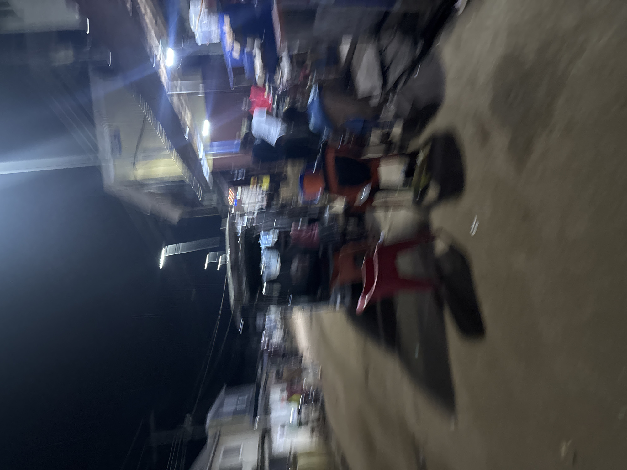 The image shows a street scene in Ihiagwa, Nigeria, at night. There are several people sitting at tables under a covered area with lights. A motorcycle is parked on the sidewalk. Buildings are visible in the background.