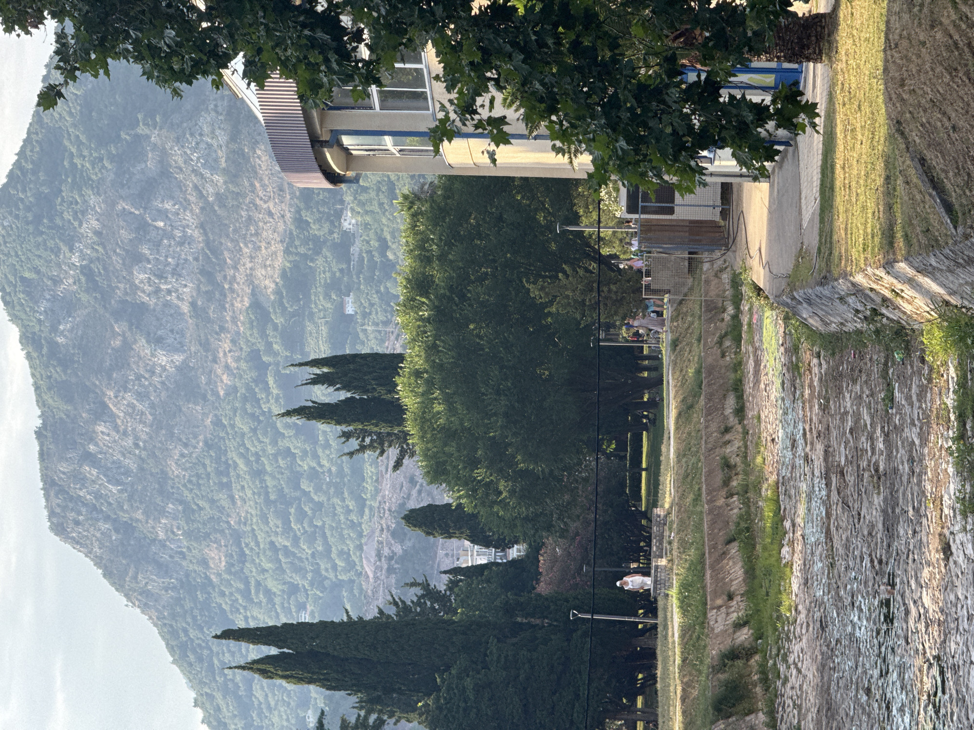 The image depicts a scenic view of a mountain range and a building, possibly in Budva, Montenegro. The mountain is covered in lush green foliage, suggesting a warm season. In the foreground, a paved path runs alongside a stone wall, with a grassy area on the other side. Several cypress trees are visible, as well as a large, leafy tree in the upper left corner. The building appears to be a modern structure with windows and a light-colored facade. There are some people in the background, and the scene suggests a peaceful, outdoor setting, possibly a park or a promenade. The lighting indicates it might be late afternoon or early evening, with the sun casting long shadows. No visible text is present.