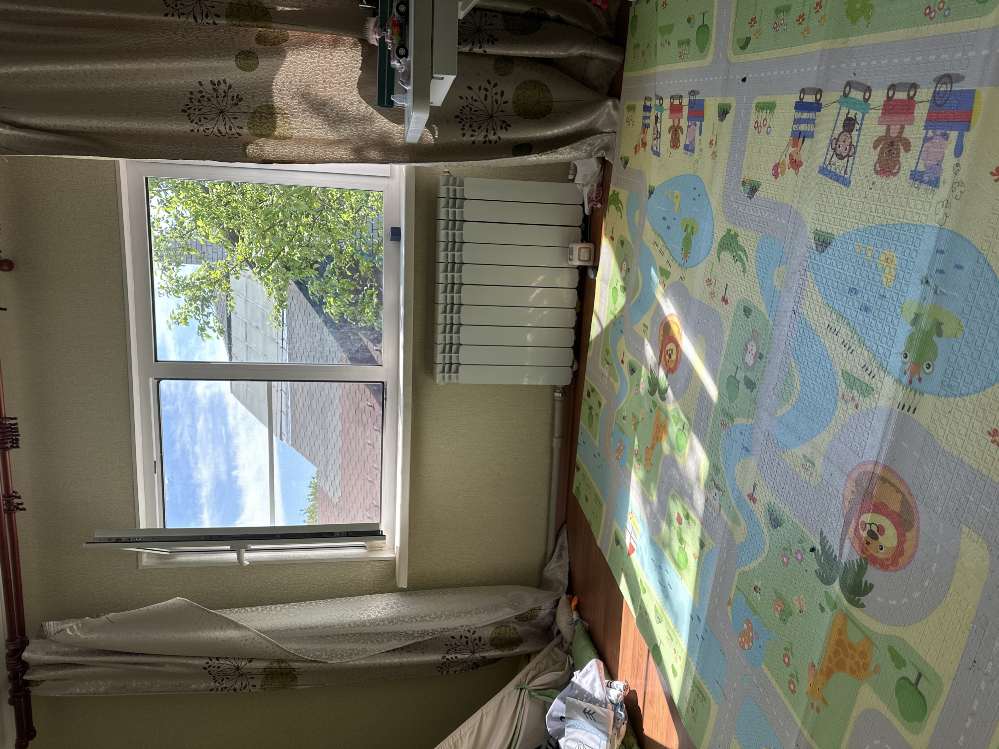 A brightly lit room in Nizhny Novgorod, Russia, contains a window showing a cloudy sky and tree leaves. A white radiator is next to the window, and a colorful children's playmat with animal and road designs is on the floor. The room also has curtains and what appears to be part of a tent or play structure in the corner.